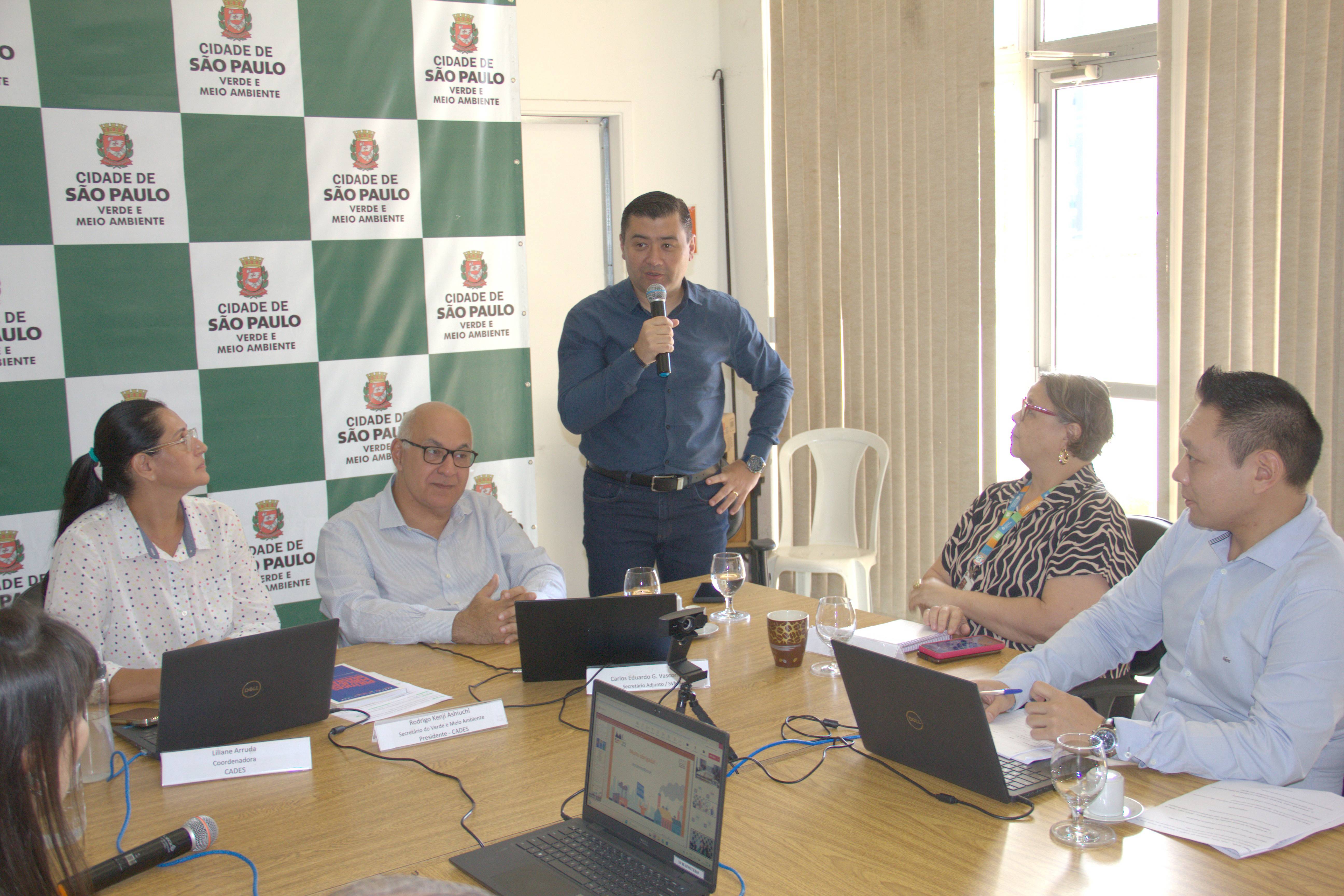 Foto do secretário Rodrigo Aschiuchi no centro, de pé, segurando um microfone enquanto discursa em frente a uma mesa de madeira. Várias pessoas estão ao redor, prestando atenção no que ele diz. Ao fundo, um banner quadriculado nas cores branco e verde escuro com o logo da Secretaria do Verde e do Meio Ambiente com os seguintes dizeres: "Cidade de São Paulo. Verde e Meio Ambiente".   