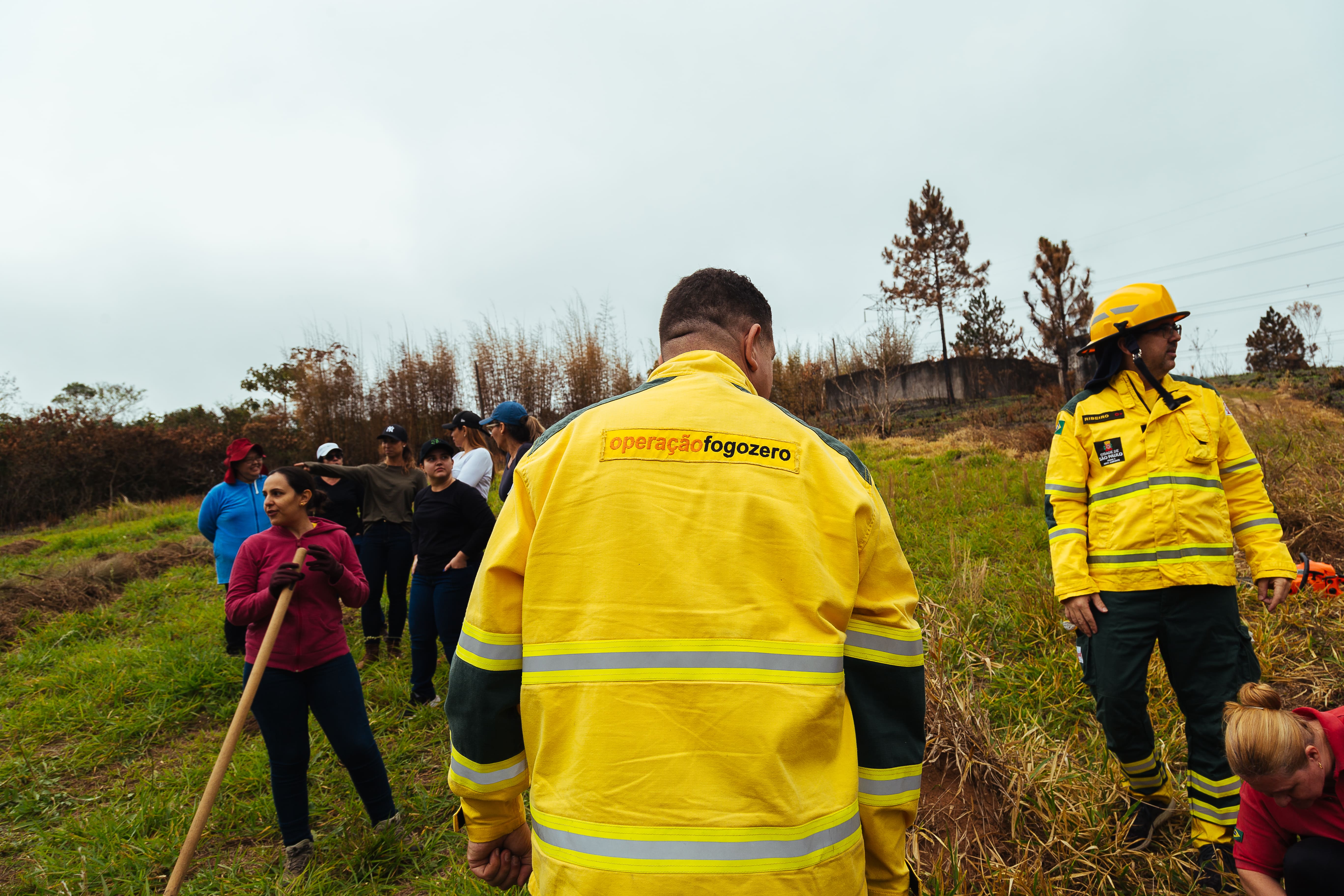 Profissional do gênero masculino utilizando uma casaco amarelo de proteção contra fogo, com os seguintes dizeres escrito nas costas "Operação Fogo Zero". Ele está de costas para câmera. Ao seu lado, há outro profissional, vestido com a mesma roupa, mas ele também usa um capacete para proteção. Do lado esquerdo da foto, há algumas pessoas juntas ao fundo. Uma mulher aponta o dedo para direção esquerda, enquanto parece conversar com mais dois homens. Na sua frente, há uma mulher segurando uma vassoura rastelo de ferro. Todos eles estão em uma área verde com gramado.   