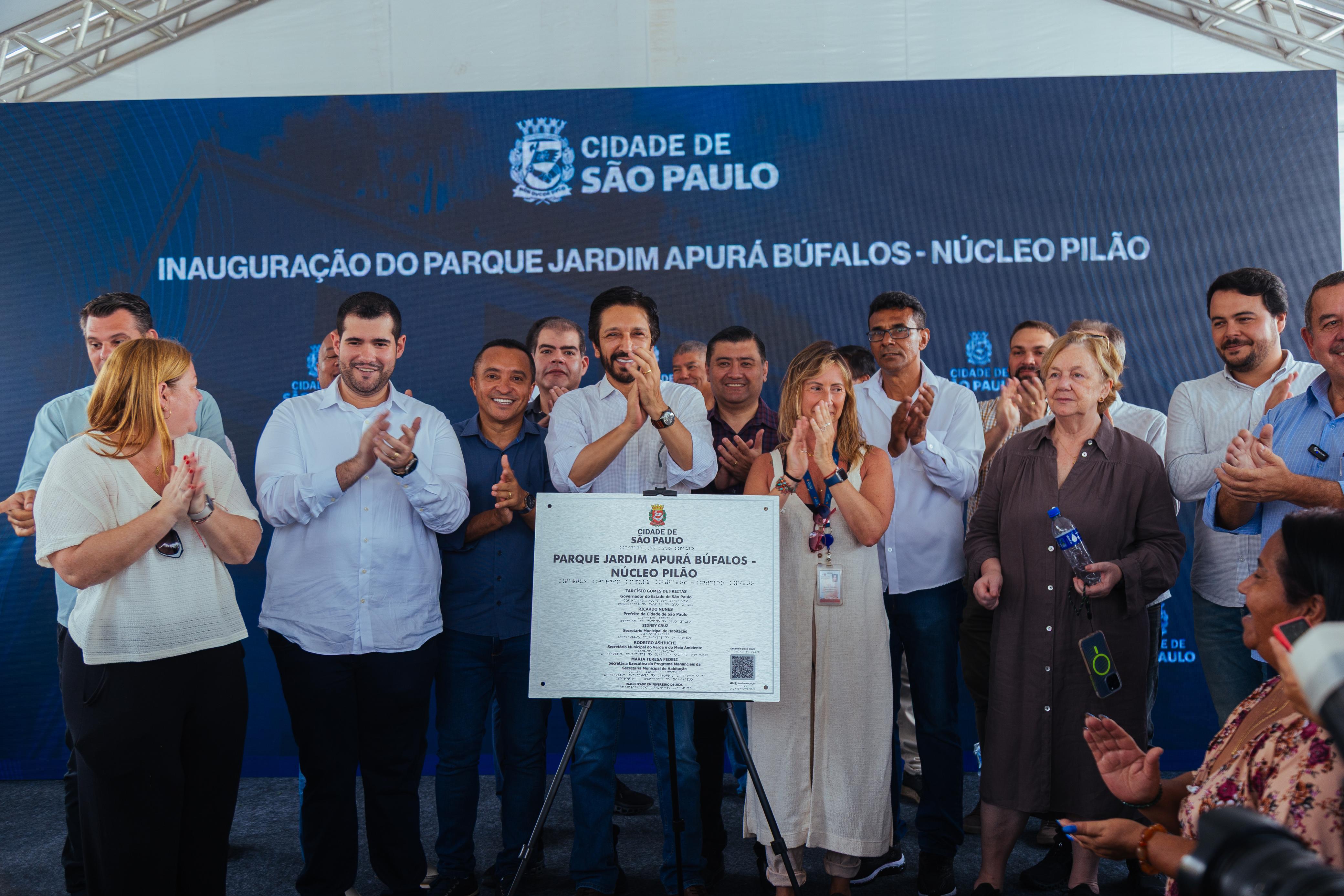 Um grupo de pessoas ao lado do prefeito Ricardo Nunes e do secretário da SVMA Rodrigo Ashiuchi, posando para uma foto em frente a um painel azul. O painel tem o texto "CIDADE DE SÃO PAULO" e "INAUGURAÇÃO DO PARQUE JARDIM APURÁ BUFALOS NÚCLEO PILÃO". As pessoas estão sorrindo e algumas aplaudem