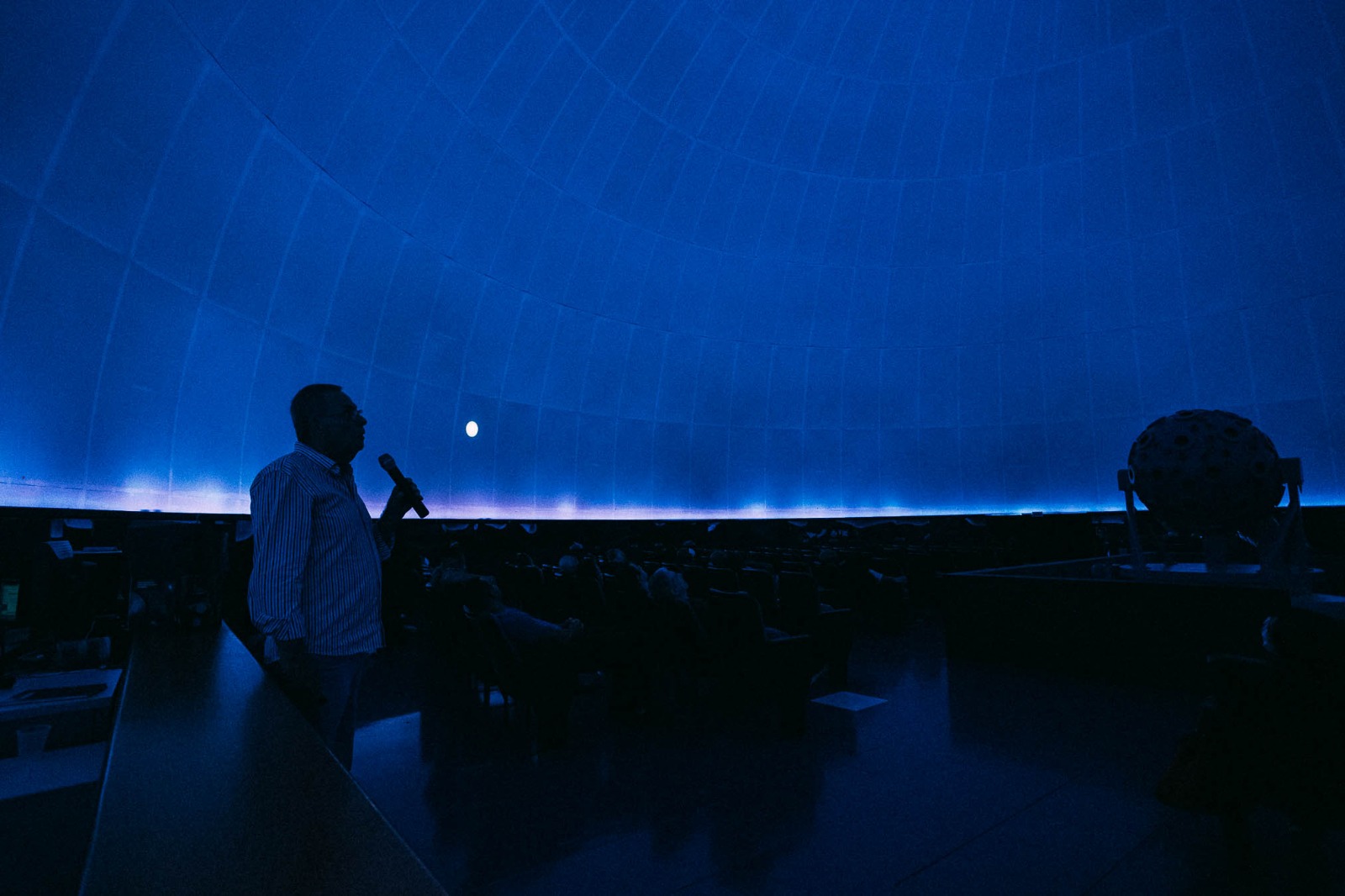 A imagem mostra o interior de um planetário com uma cúpula iluminada em tons de azul. No lado esquerdo, um homem segura um microfone e parece estar fazendo uma apresentação para um público sentado em cadeiras dispostas em um formato circular. No lado direito, há um projetor esférico, possivelmente responsável pela exibição do céu simulado na cúpula. A iluminação cria um ambiente imersivo, destacando a projeção de um corpo celeste, que pode ser a Lua, na superfície interna da cúpula. A cena transmite uma atmosfera educativa e envolvente, típica de apresentações astronômicas.