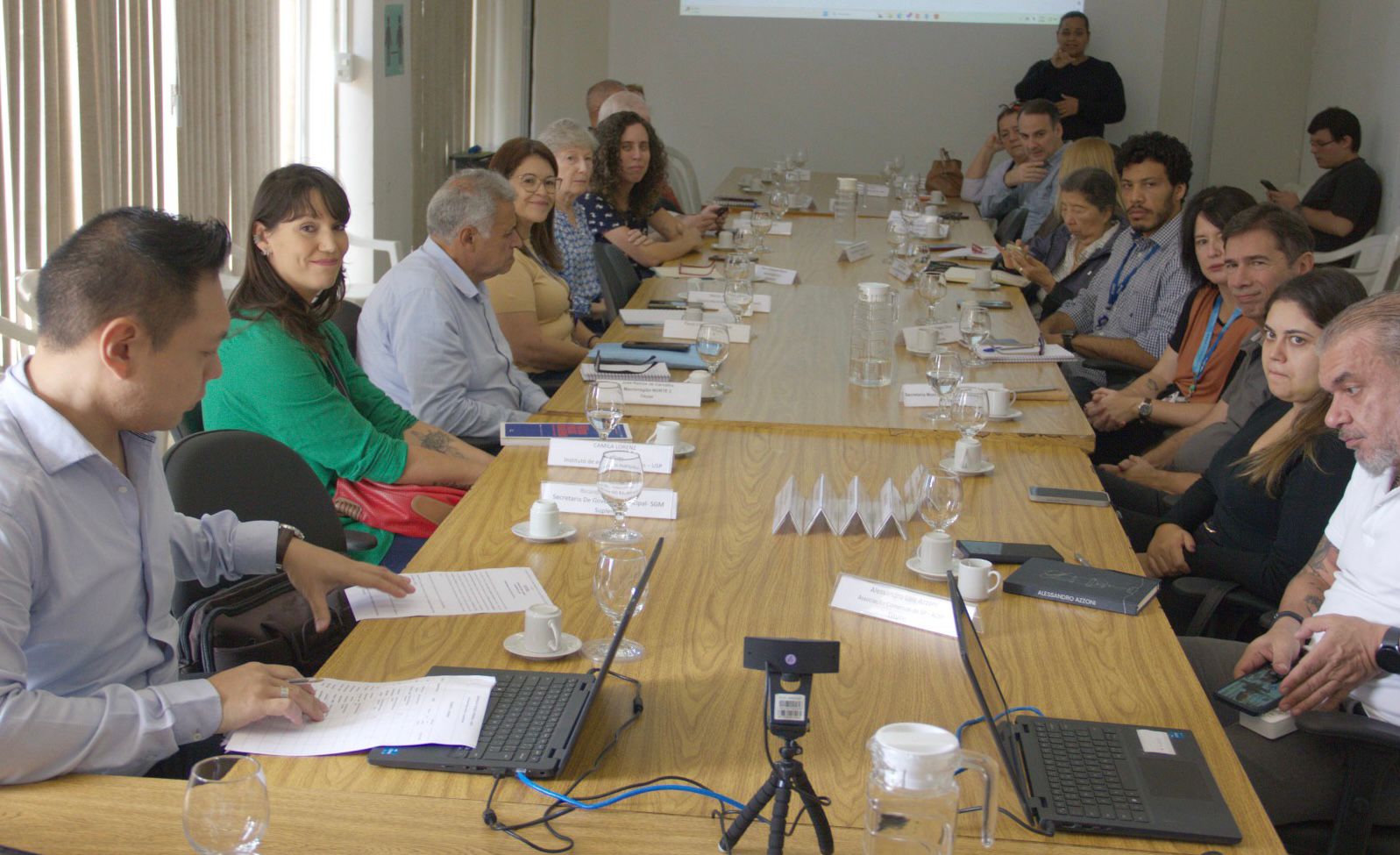 Várias pessoas sentadas em frente a uma mesa de madeira, durante a reunião do CADES Municipal. Na mesa, há notebooks, cadernos e folhas de papel para anotação, livros e taças com água. Todos olham para frente, em direção a câmera. 