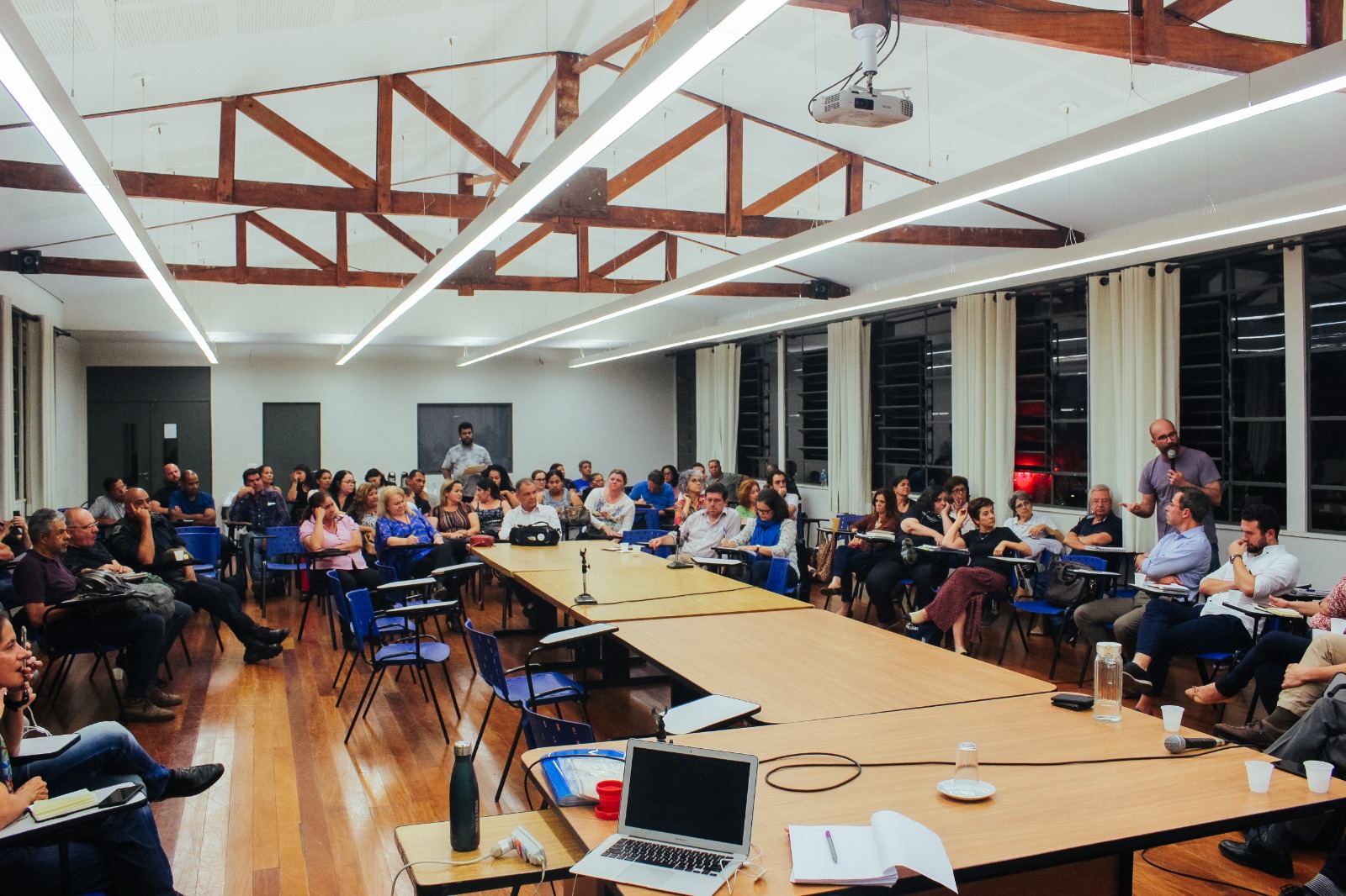 A imagem mostra uma sala ampla e bem iluminada, onde ocorre uma reunião ou audiência pública com muitas pessoas sentadas em cadeiras azuis. No centro há mesas retangulares de madeira clara dispostas em formato de "U". As paredes laterais possuem grandes janelas com cortinas brancas. O teto é branco com estruturas de madeira aparente em forma de “V” invertido e iluminação de trilhos com luzes brancas lineares.  As pessoas estão vestindo roupas de cores variadas, incluindo azul, branco, rosa, roxo, preto e cinza. À direita da imagem, um homem careca de camiseta roxa está em pé, falando com um microfone, enquanto outros o observam atentamente. Sobre a mesa central há notebooks, uma garrafa de água, copos plásticos brancos, microfones e papéis. O chão é de madeira envernizada em tom marrom claro. Ao fundo, um projetor está preso ao teto. A atmosfera é de concentração e participação ativa.