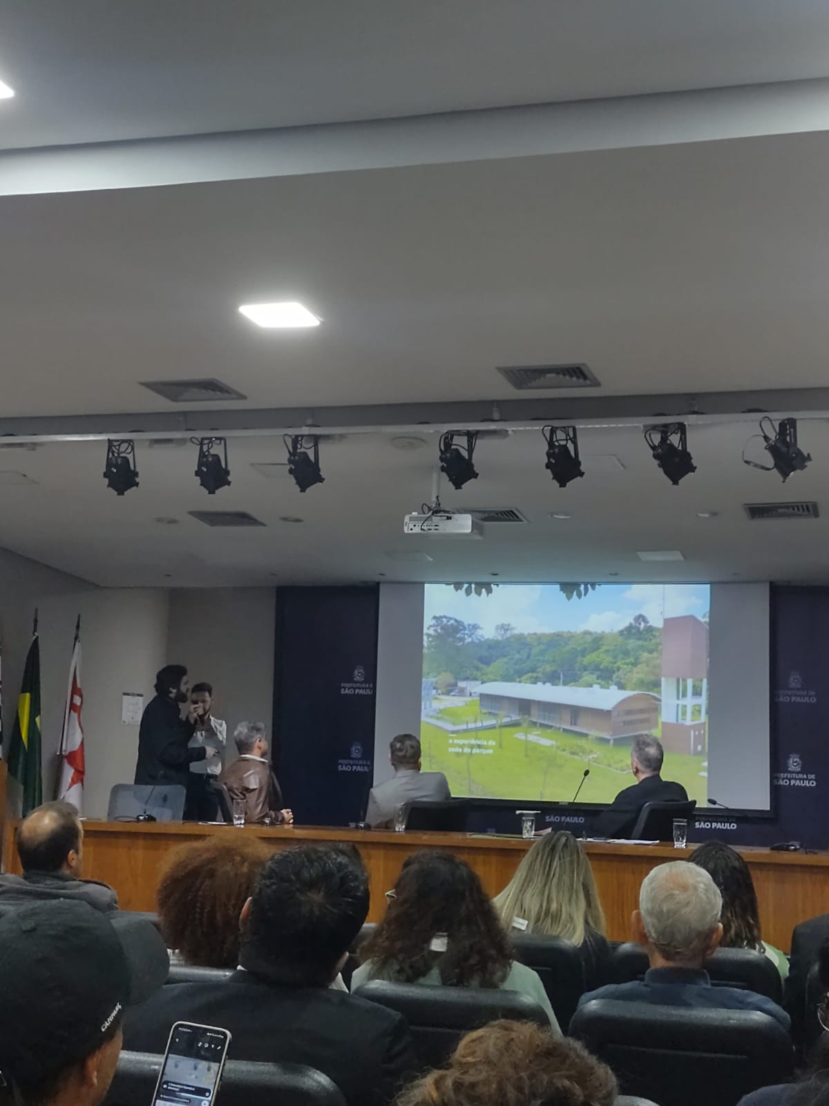 O ChatGPT disse: A imagem mostra um auditório durante uma apresentação institucional, provavelmente de um órgão público da cidade de São Paulo, como indicado pelas bandeiras oficiais ao fundo e pelos painéis com o brasão e o nome da prefeitura. No centro da cena, duas pessoas estão de pé com microfone, enquanto outras assistem à apresentação projetada, que exibe a imagem de uma edificação com áreas verdes ao redor e os dizeres “a importância da sede do parque”. O público está sentado, atento, e muitos tiram fotos ou gravam com seus celulares. A atmosfera é formal, típica de eventos técnicos, audiências ou reuniões públicas.