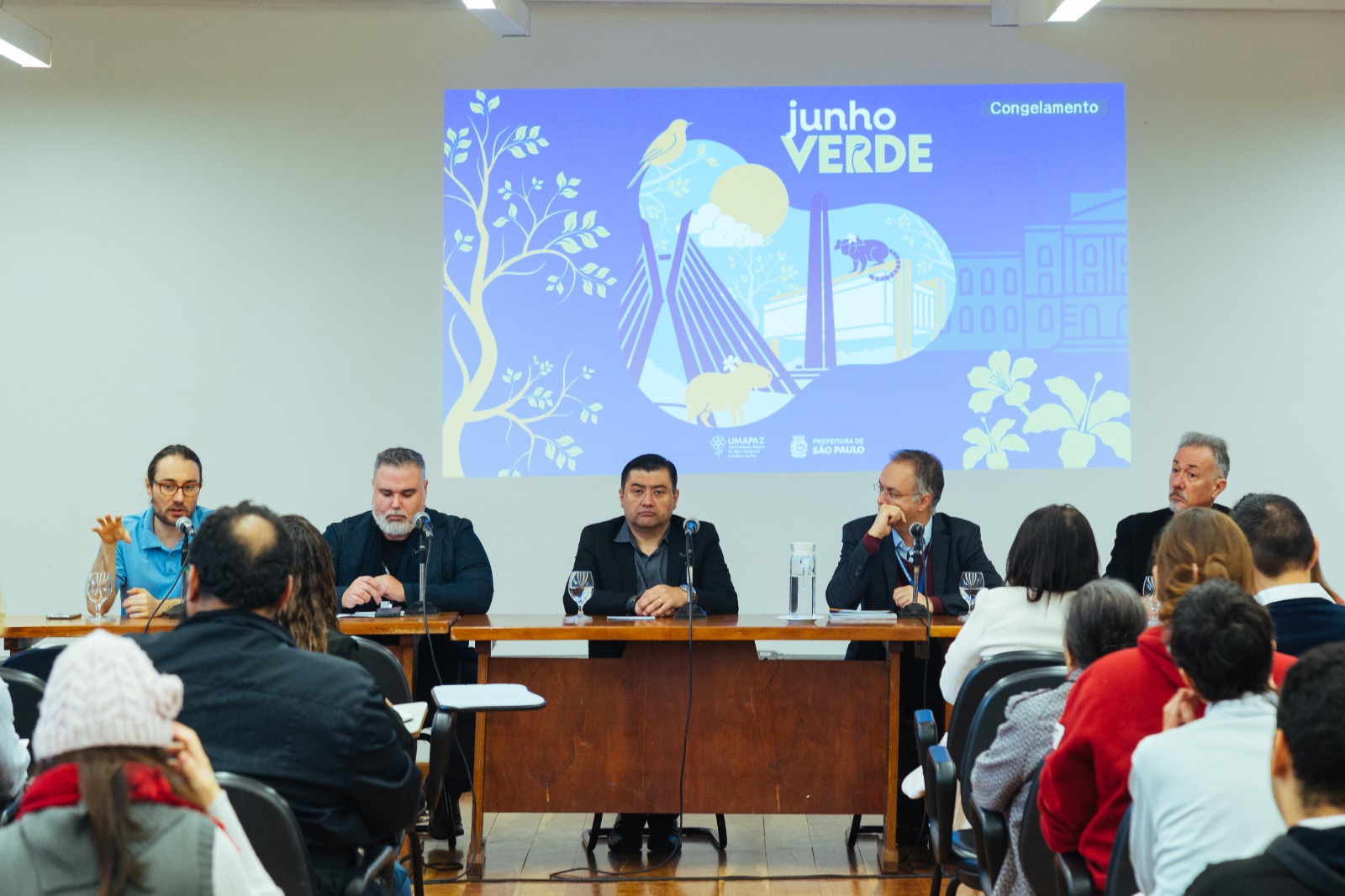 A imagem mostra um painel da programação do Junho Verde, com foco no tema “Congelamento”, promovido pela Prefeitura de São Paulo por meio da UMAPAZ (Universidade Aberta do Meio Ambiente e da Cultura de Paz). O evento ocorre em um auditório, com cinco homens sentados à mesa de debate, diante de um público atento.  Ao fundo, o telão exibe uma arte gráfica com elementos ambientais e urbanos, incluindo árvores, flores, animais e a ponte estaiada — símbolo da cidade de São Paulo — reforçando a proposta de reflexão sobre sustentabilidade, proteção ambiental e planejamento urbano. A presença de microfones e copos d'água indica uma mesa-redonda ou palestra com caráter institucional e técnico.  Se desejar, posso sugerir uma legenda ou linha-fina jornalística para essa imagem.