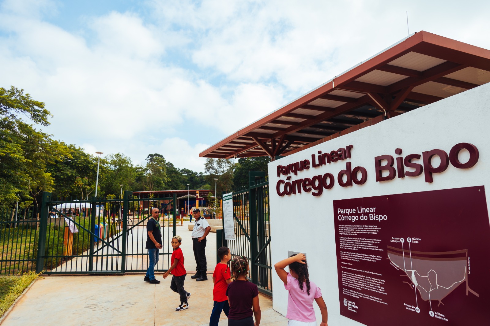 A imagem mostra a entrada principal do Parque Linear Córrego do Bispo, localizado na cidade de São Paulo. A entrada tem um letreiro em alto-relevo com o nome do parque e um painel informativo com mapa e orientações sobre o espaço. O projeto urbanístico se caracteriza por trilhas naturais, núcleos de lazer, áreas verdes e um traçado que acompanha o curso do córrego, como é típico dos parques lineares.  Na cena, algumas crianças e adultos interagem na entrada, sugerindo que o local é voltado ao uso comunitário e familiar. O visual moderno da infraestrutura, com portões largos, cobertura metálica e sinalização clara, reforça o investimento em equipamentos públicos de qualidade nas periferias urbanas.  O parque é uma iniciativa da Cidade de São Paulo, por meio da Secretaria do Verde e do Meio Ambiente, como indicado nos elementos gráficos da placa. Ele cumpre papel importante na promoção da sustentabilidade urbana, contenção de enchentes e oferta de áreas de lazer em regiões carentes de infraestrutura verde.