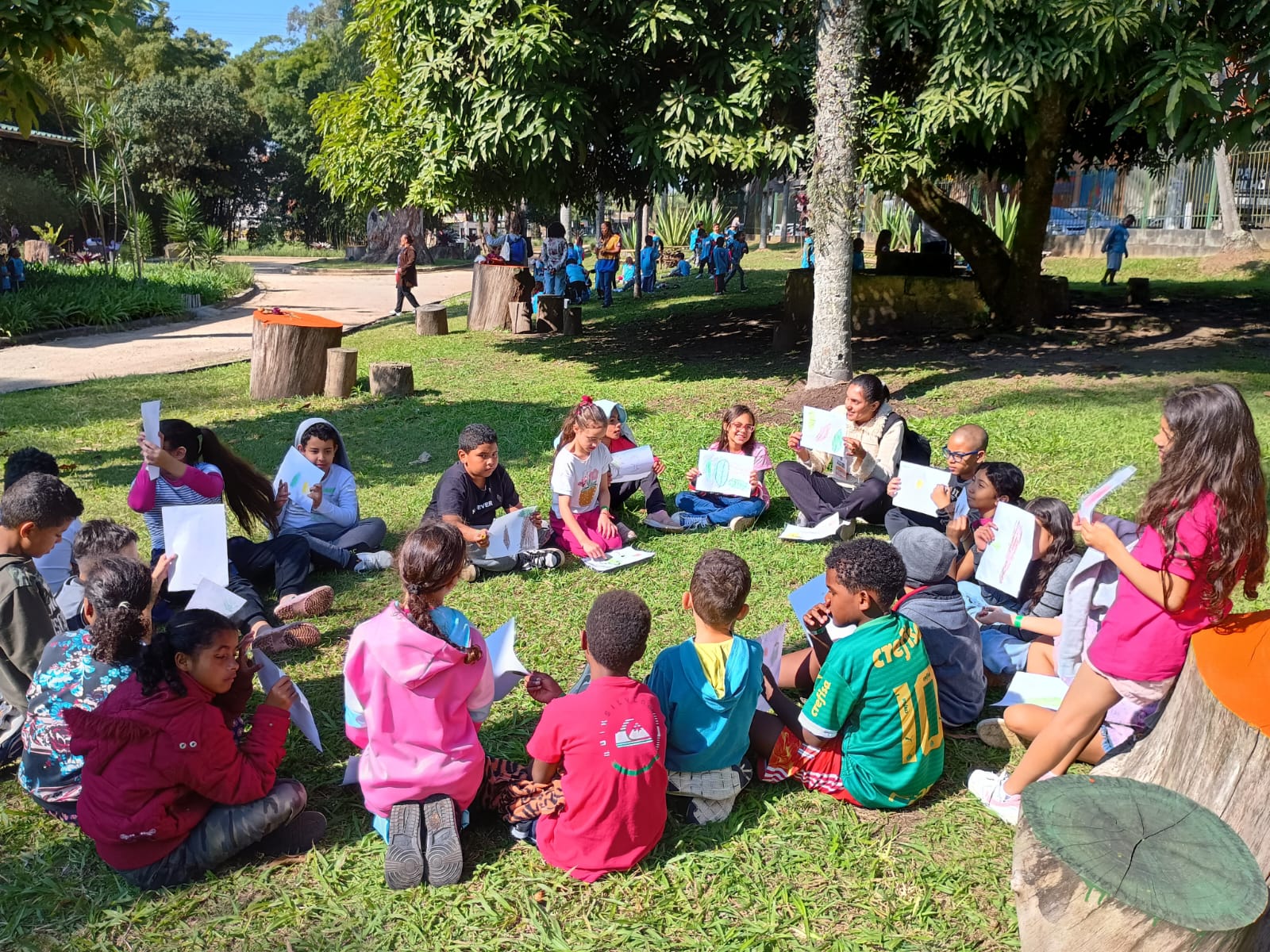 Cerca de 25 crianças estão sentadas em roda sobre a grama em um parque arborizado, participando de uma atividade ao ar livre. Algumas seguram folhas de papel com desenhos coloridos, enquanto uma mediadora interage com o grupo. Ao fundo, é possível ver outras crianças e adultos caminhando, além de bancos de troncos e árvores que oferecem sombra. O clima é de aprendizado e recreação.