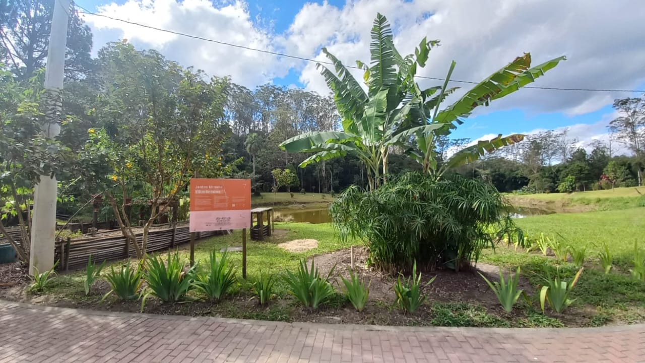 Vista de um jardim agroecológico com vegetação diversa, incluindo bananeiras, arbustos e plantas ornamentais. Ao centro, há uma placa laranja com os dizeres “Jardins filtrantes: Vilson de Souza”, indicando um sistema de tratamento natural de água. Ao fundo, vê-se um pequeno lago cercado por áreas verdes e árvores altas. O céu está parcialmente nublado com nuvens brancas e céu azul.