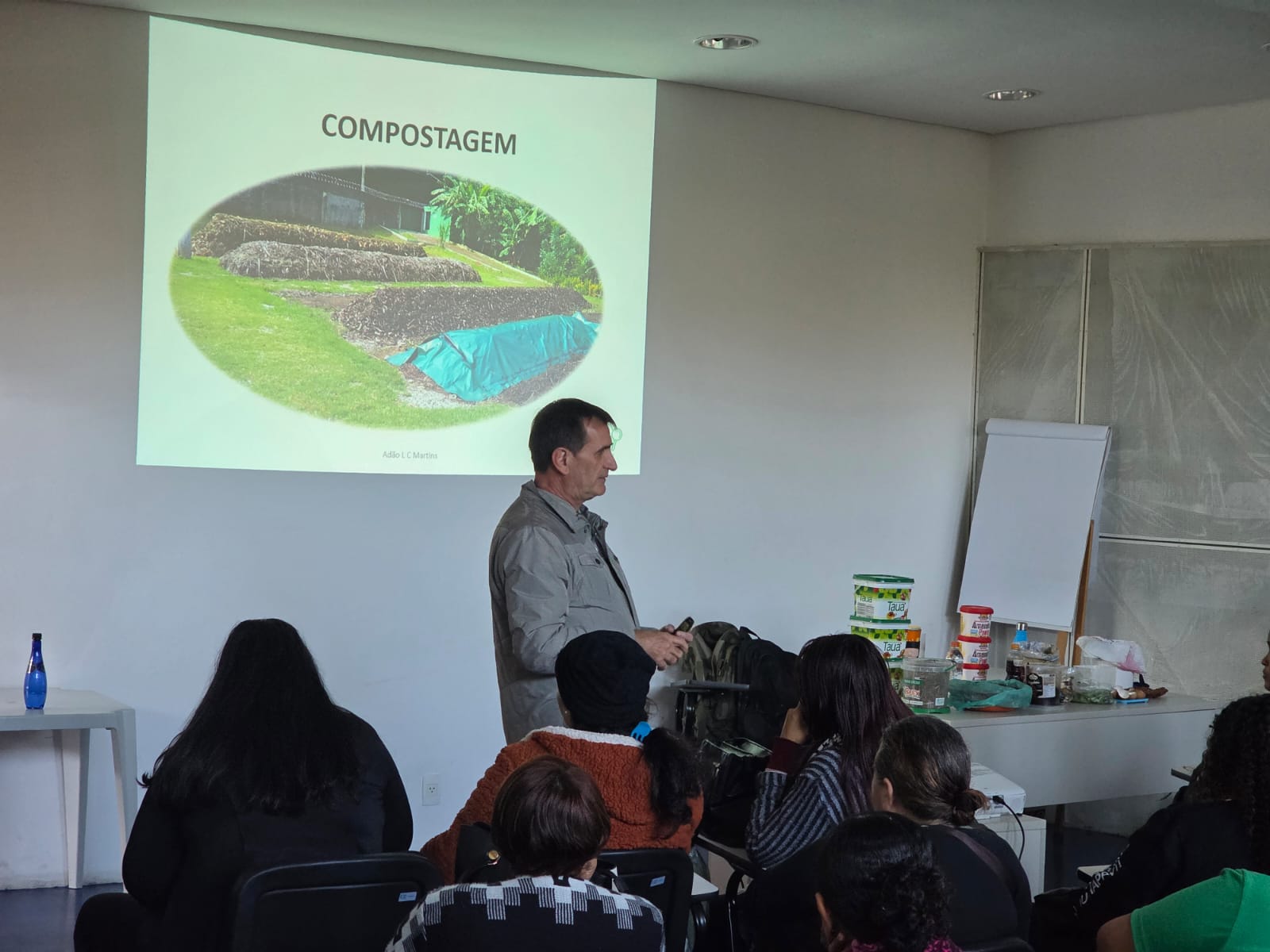 Homem ministra palestra sobre compostagem em uma sala com público atento. Ao fundo, há uma projeção com a palavra "COMPOSTAGEM" e uma imagem de leiras de compostagem cobertas por lona. Em primeiro plano, estão participantes sentados. À direita, uma mesa com materiais de apoio, incluindo potes reutilizados, e um quadro branco de apoio encostado na parede.