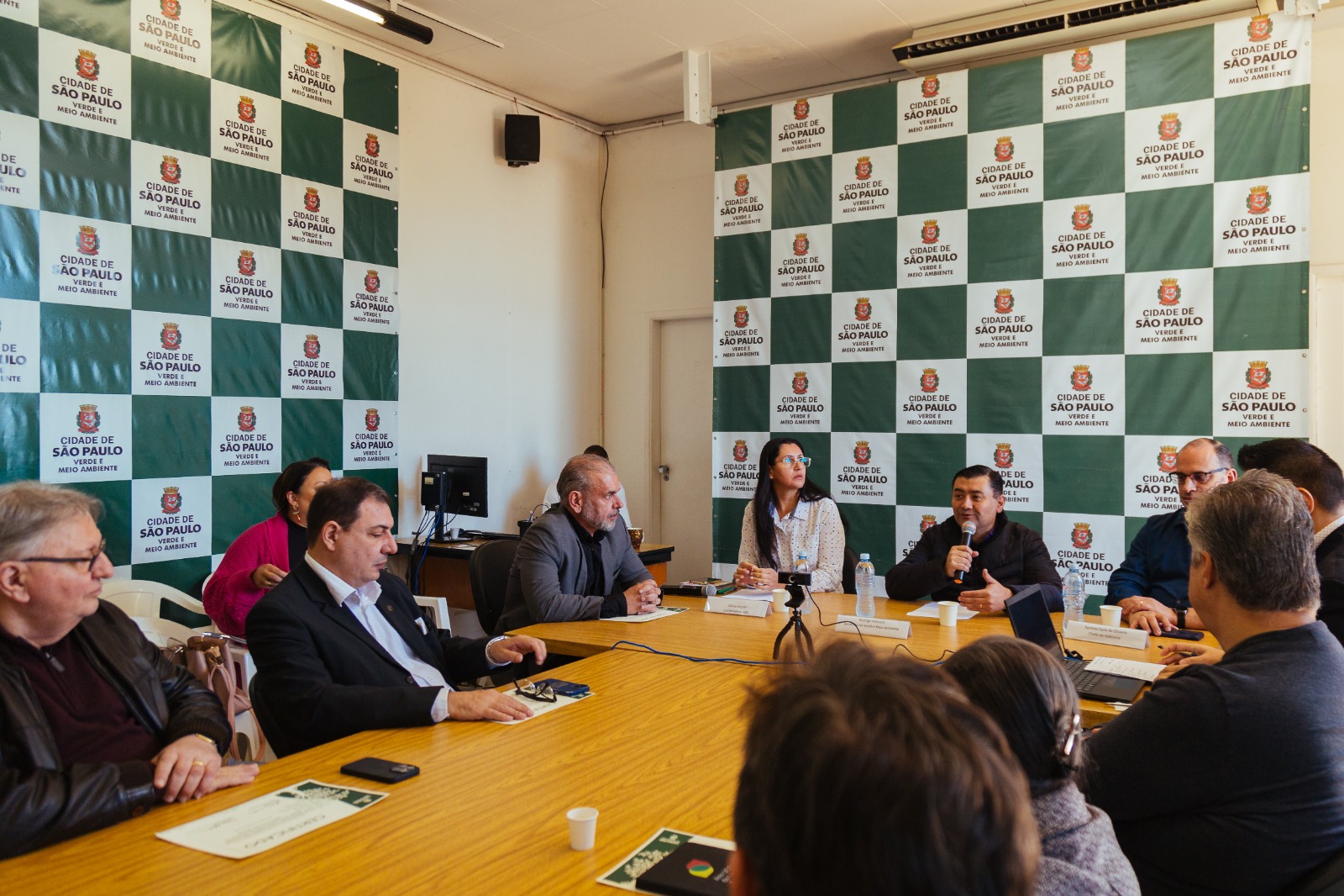 A imagem mostra uma reunião realizada em uma sala institucional da Prefeitura de São Paulo, com destaque para o fundo decorado com o logotipo da "Cidade de São Paulo – Verde e Meio Ambiente". Sentados ao redor de uma mesa retangular de madeira clara, diversos participantes, entre eles gestores públicos, técnicos e convidados, participam de uma discussão. Um dos homens, segurando o microfone, parece conduzir ou responder uma fala, sugerindo que a reunião tem caráter técnico ou deliberativo. Há microfones, câmeras e material impresso sobre a mesa, o que indica um encontro formal, possivelmente relacionado à gestão ambiental, planejamento urbano ou apresentação de projetos da Secretaria do Verde e do Meio Ambiente. A presença de computadores ao fundo e materiais oficiais reforçam o contexto administrativo e institucional da ocasião.