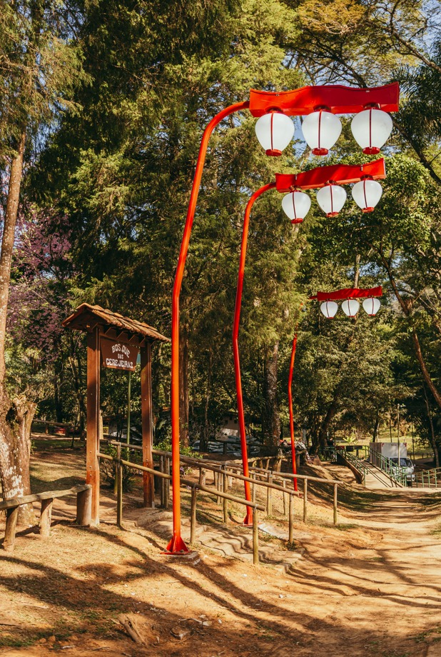 A imagem mostra a entrada do Bosque das Cerejeiras, localizado no Parque do Carmo, na zona leste de São Paulo. O espaço é caracterizado pela presença de luminárias com design típico japonês e por uma arborização densa, com destaque para as cerejeiras, que florescem entre julho e agosto. O local é um dos principais pontos da cidade para contemplação da florada das sakuras, símbolo da cultura japonesa e da renovação da natureza. O Parque do Carmo é também sede do tradicional Festival das Cerejeiras, evento que celebra a cultura nipônica e atrai milhares de visitantes todos os anos.