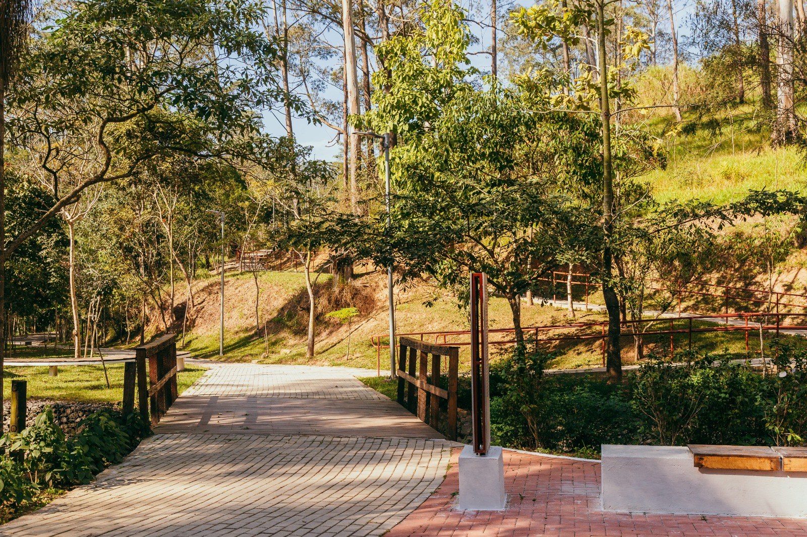 Caminho pavimentado com blocos intertravados atravessa uma ponte de madeira em meio a uma área verde com árvores e vegetação nativa. À direita, há um banco de madeira sobre base de concreto e um totem moderno em metal. O cenário é de um parque com relevo variado, trilhas, gramado e postes de iluminação, em um dia ensolarado.