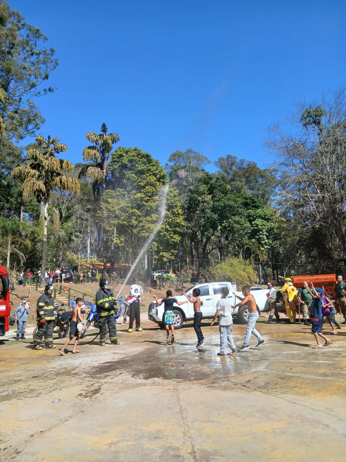 Crianças brincam sob o jato d’água de uma mangueira controlada por bombeiros uniformizados em um parque. A cena ocorre em um dia ensolarado, com muitas árvores ao fundo, e o ambiente é descontraído com a presença de adultos e mascotes observando.