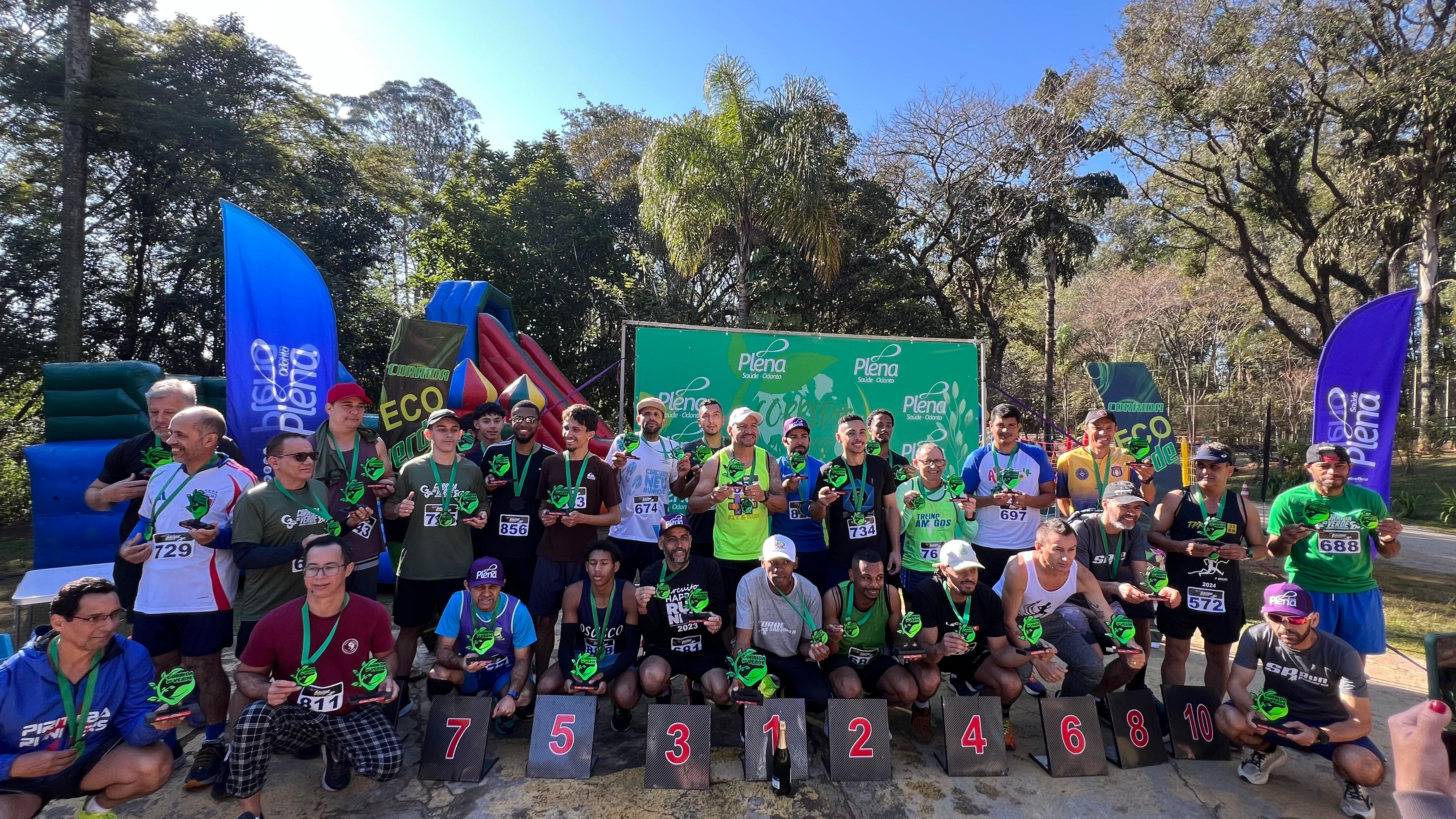 Grupo de corredores posa para foto coletiva após premiação da “Corrida Eco Plena”. Os participantes estão com medalhas no pescoço e troféus verdes nas mãos, posicionados atrás de placas numeradas de 1 a 10. Ao fundo, há infláveis coloridos, árvores e bandeiras com a marca do evento.