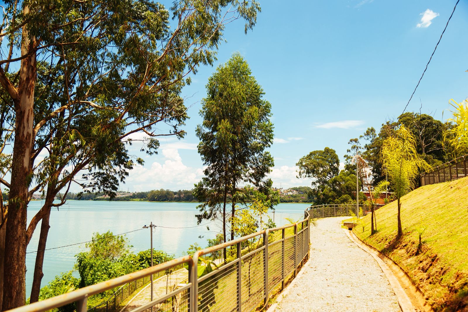 Caminho de pedrinhas cercado por gradil metálico contorna um trecho arborizado à beira de uma represa, com vegetação variada e árvores altas. O céu azul claro e o reflexo da água completam a paisagem serena em um dia ensolarado. À direita, o terreno gramado inclinado acompanha o traçado do caminho.