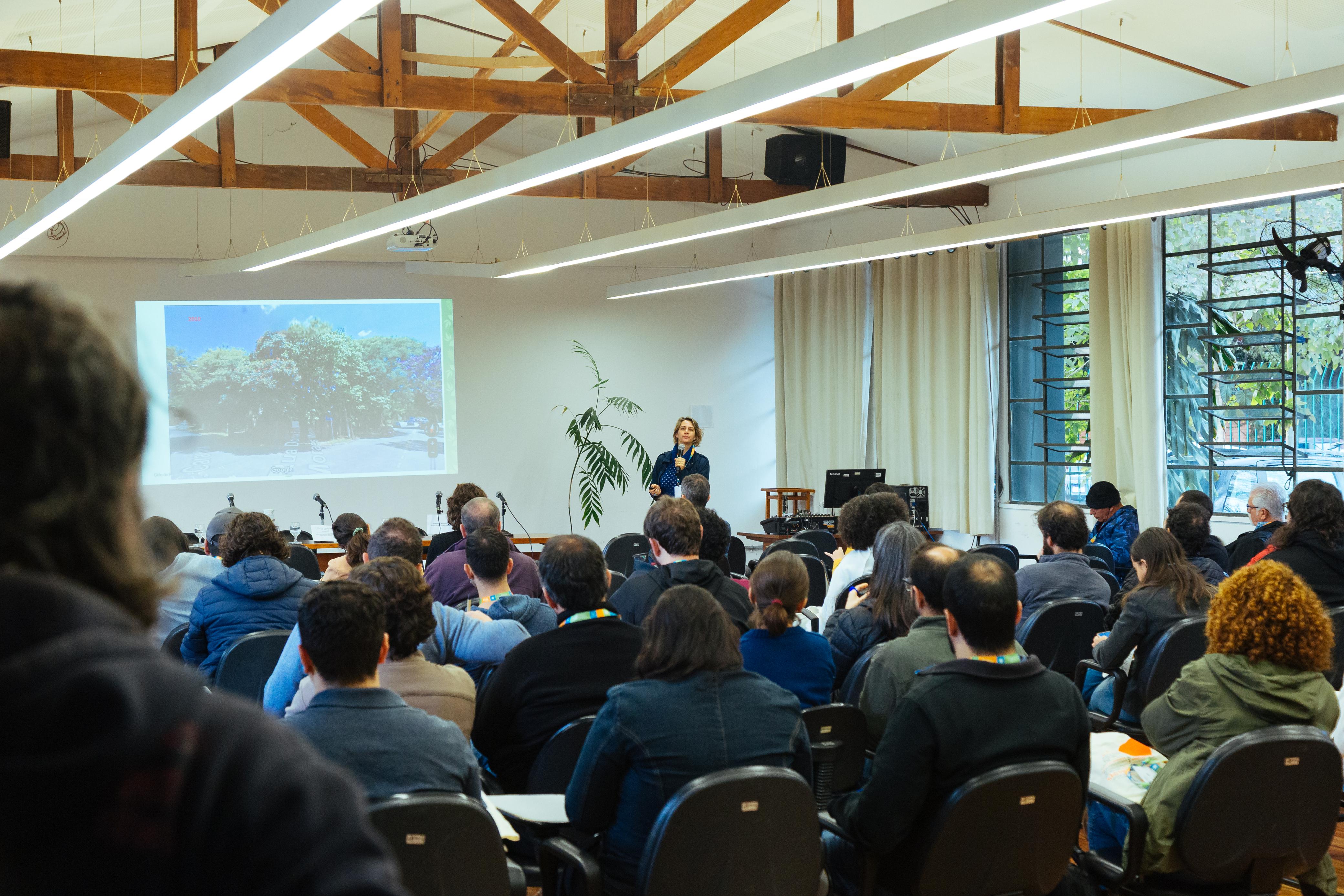Auditório com diversas pessoas sentadas assistindo a uma palestra. Uma mulher está em pé à frente, apresentando uma imagem projetada de uma rua arborizada. O ambiente tem estrutura de madeira no teto, janelas grandes ao fundo e plantas decorativas. O público parece atento à apresentação, que integra um ciclo de palestras sobre arborização urbana.