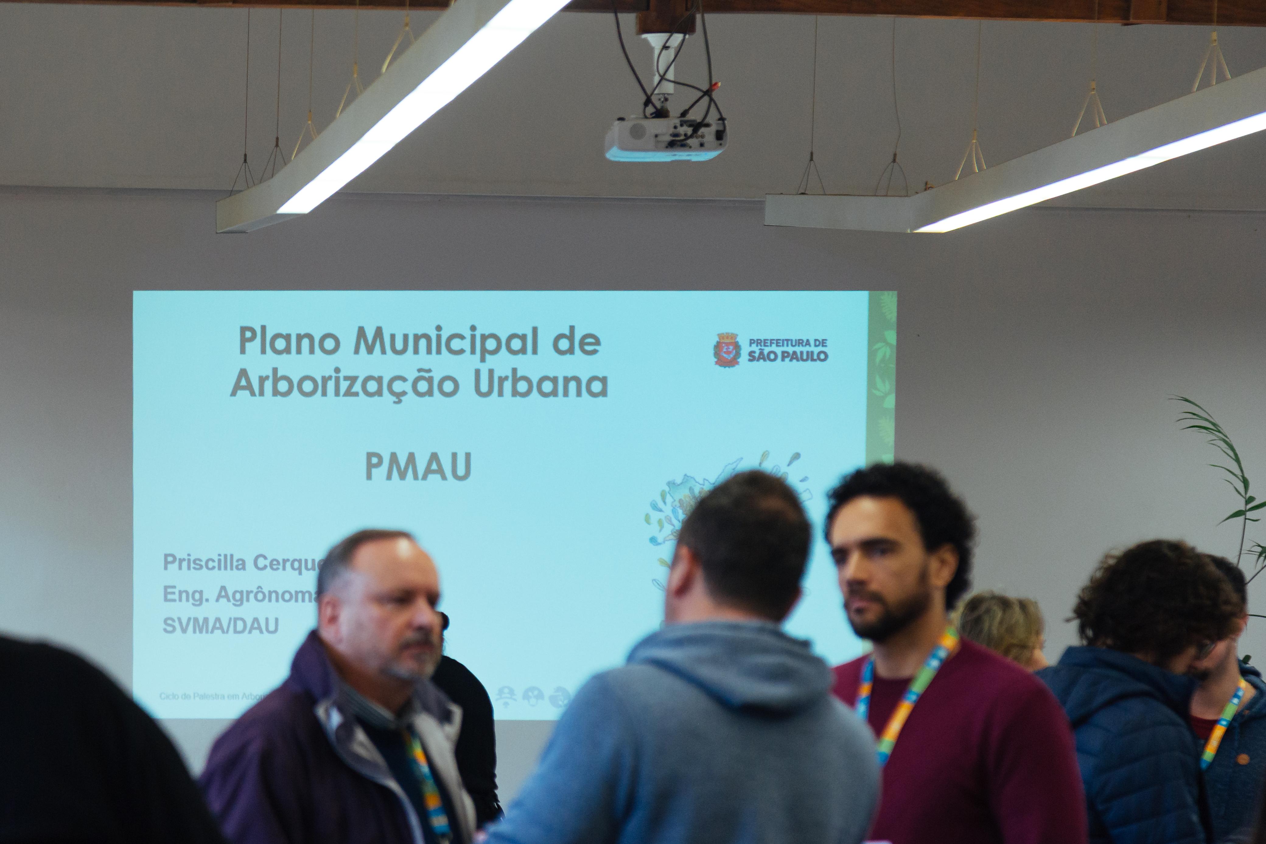 Sala de eventos com algumas pessoas em pé conversando em frente a uma tela de projeção. A projeção exibe a apresentação intitulada “Plano Municipal de Arborização Urbana – PMAU”, com o nome da engenheira agrônoma Priscilla Cerqueira e a sigla SVMA/DAU. No canto superior direito da tela está o logotipo da Prefeitura de São Paulo. A imagem mostra parte de um evento ou seminário relacionado ao tema de arborização urbana.