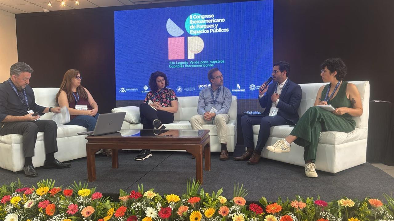 A imagem mostra seis pessoas sentadas em dois sofás brancos sobre um palco, participando de um painel de discussão. No fundo, há um grande telão azul com o logotipo e o nome do evento em letras brancas e coloridas: “I Congreso Iberoamericano de Parques y Espacios Públicos”, acompanhado do subtítulo em verde “Un legado verde para nuestras Capitales Iberoamericanas”.  As cores do logotipo são vibrantes: azul-claro, verde-claro, rosa e bege, formando formas geométricas. O chão do palco é preto e, à frente, há um arranjo de flores coloridas, com tons vivos de amarelo, vermelho, laranja, rosa e branco, contrastando com o verde das folhas.  As roupas das pessoas variam entre cores neutras e escuras (preto, cinza, azul-marinho) e cores mais vivas, como verde e estampas coloridas. Uma mesa de centro marrom-escura com tampo de vidro está posicionada entre os sofás.