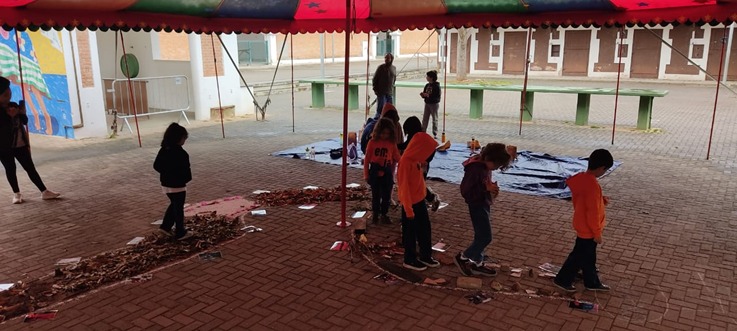 A segunda imagem retrata um grupo de crianças em um espaço externo coberto por um toldo listrado nas cores vermelho, laranja e azul. Elas percorrem um caminho no chão delimitado por folhas secas e materiais recicláveis, como garrafas e embalagens. Algumas crianças usam casacos laranja e outras roupas escuras. Ao fundo, vê-se uma área de pátio com bancos verdes e construções de tijolos aparentes.