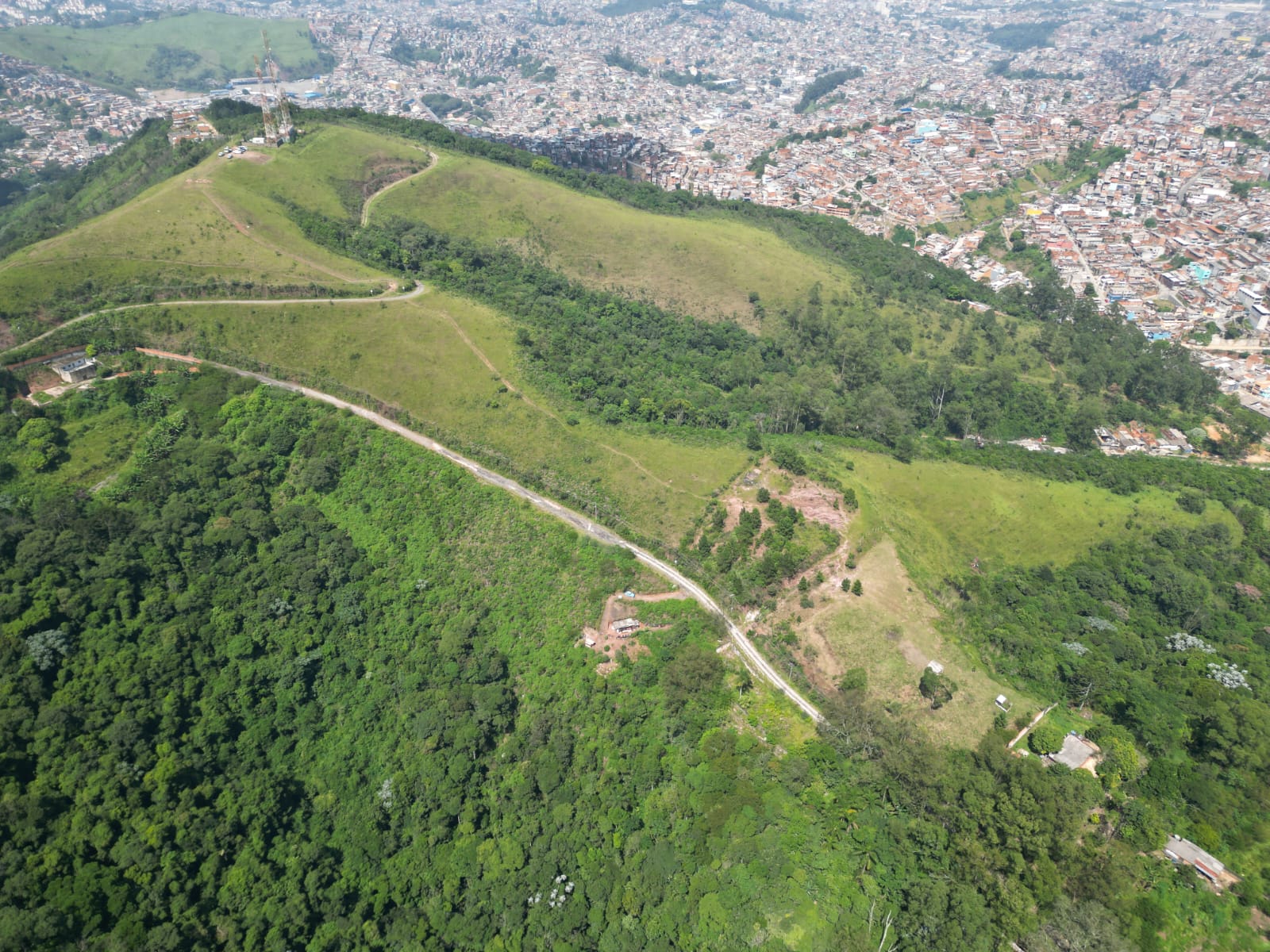 A imagem mostra uma vista aérea de uma paisagem montanhosa, com áreas verdes e uma cidade densamente povoada ao fundo. A montanha é coberta por vegetação variada, com áreas de pastagem e florestas densas. Trilhas sinuosas cortam a encosta. No topo da montanha, há uma estrutura que parece ser uma torre ou monumento. A cidade ao fundo é densa, com casas pequenas e próximas umas das outras. As cores predominantes são o verde da vegetação e tons de cinza e marrom na área urbana.
