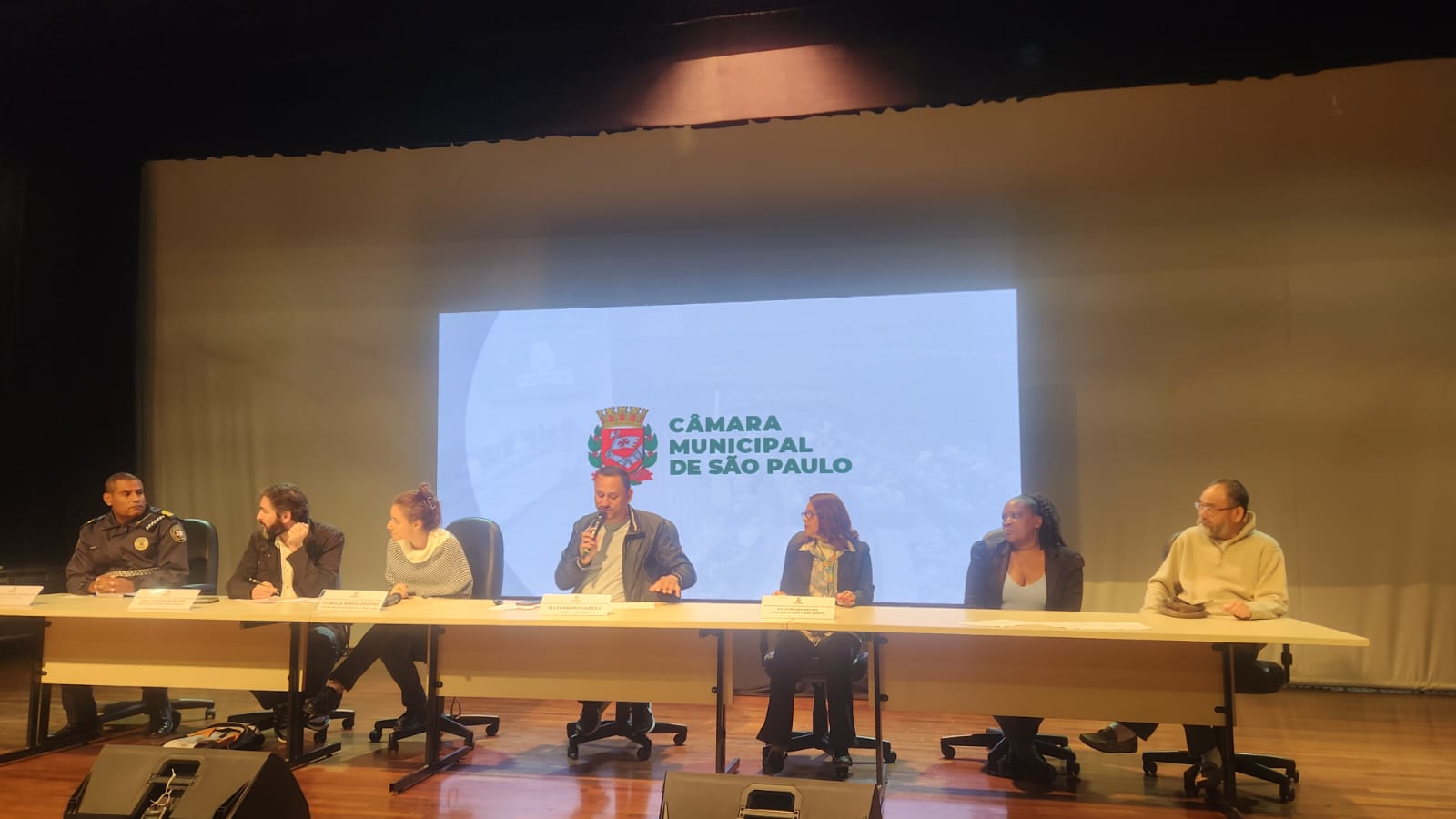 A imagem mostra uma mesa de debate com sete pessoas diversas, incluindo mulheres, homens e um agente da GCM, participando de um evento oficial na Câmara Municipal de São Paulo. Eles estão sentados lado a lado, em um ambiente acessível e acolhedor, promovendo diálogo e participação cidadã.