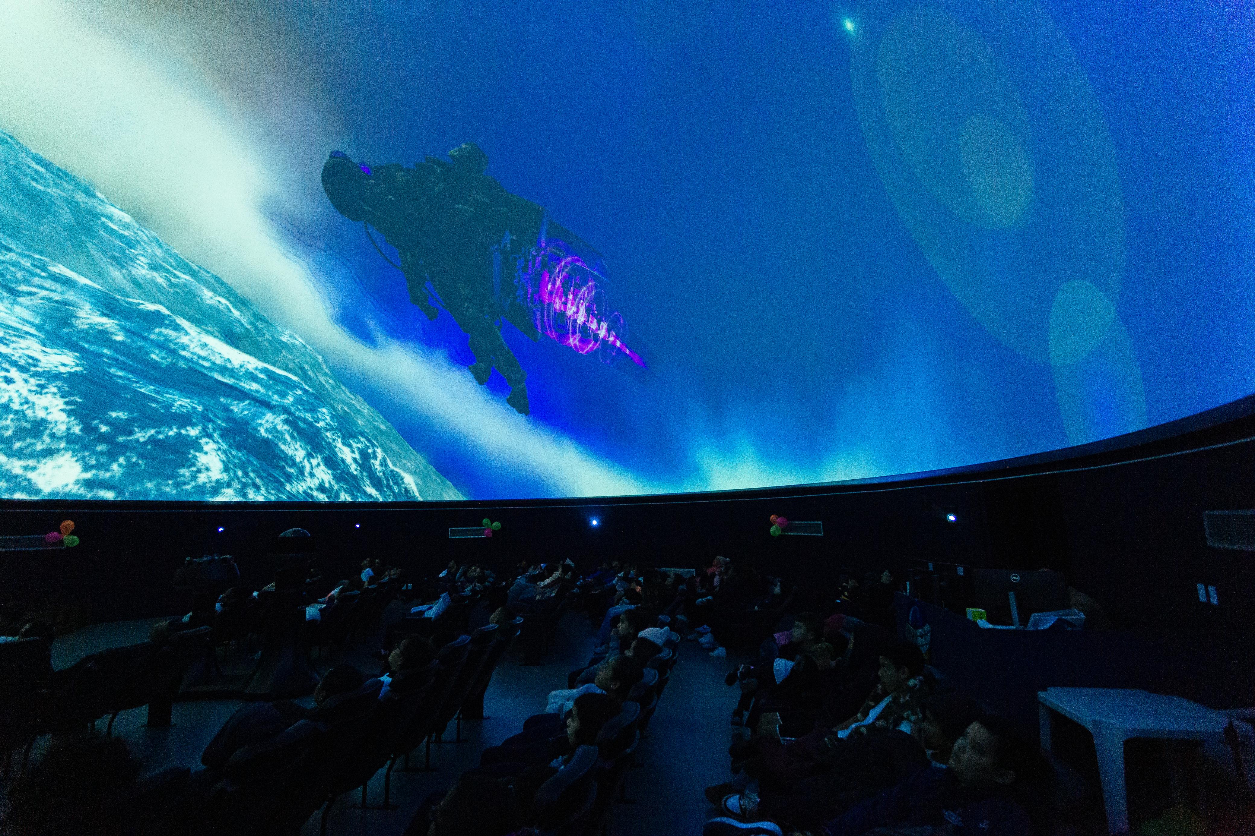 Interior de um planetário com diversas pessoas sentadas em poltronas reclinadas, assistindo a uma projeção no teto em formato de cúpula. A imagem projetada mostra a Terra vista do espaço e uma figura futurista em roxo e preto, semelhante a uma nave ou equipamento espacial.