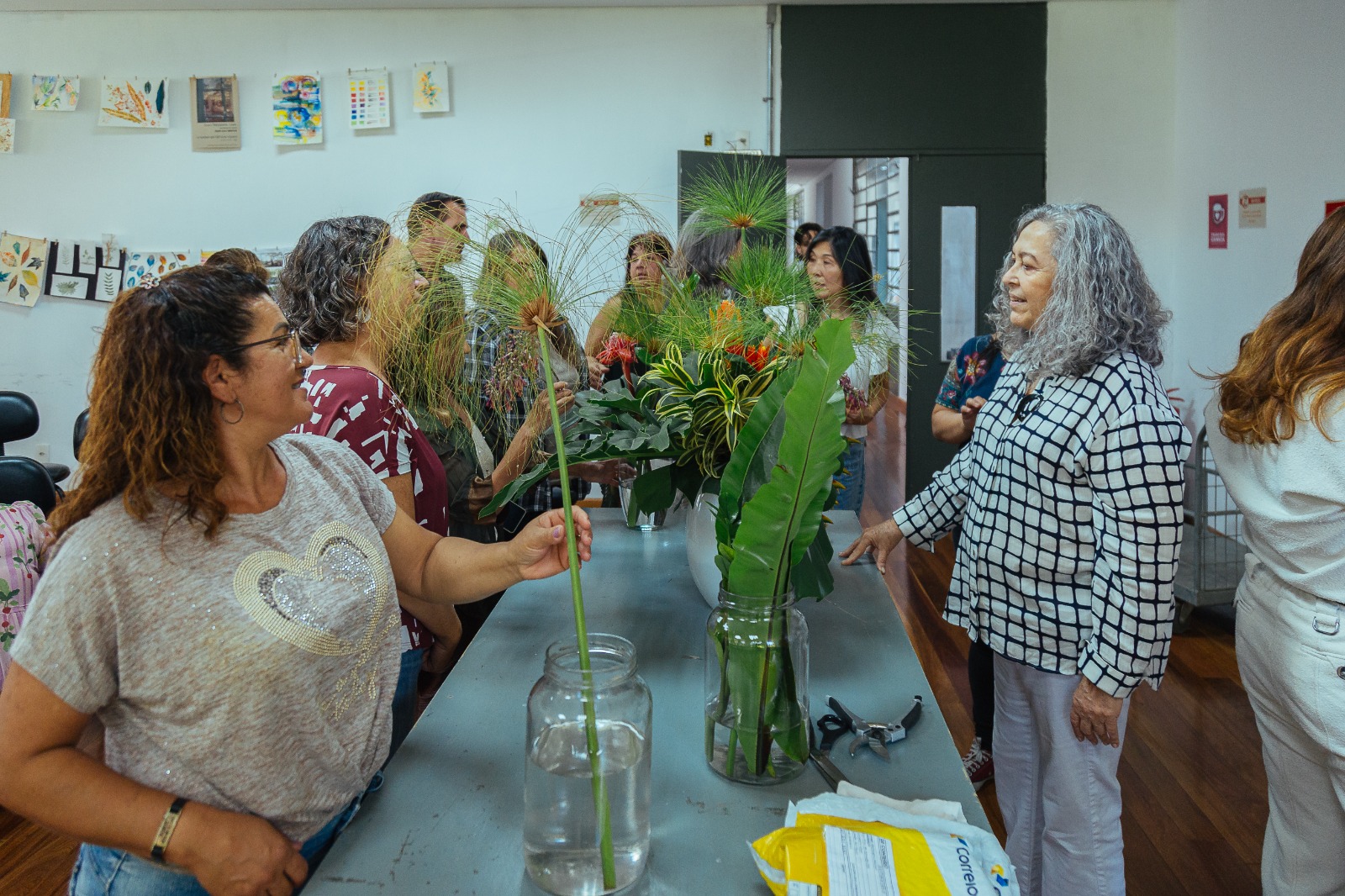 Grupo de pessoas reunidas em uma sala de aula, em torno de uma mesa com arranjos de plantas e flores em vasos de vidro. Uma mulher de cabelos grisalhos fala com os participantes, enquanto outra segura um caule comprido. Ao fundo, há desenhos e pinturas pendurados na parede.