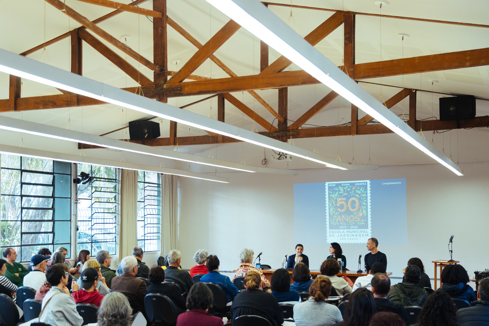 Auditório com diversas pessoas sentadas assistindo a uma mesa de debate composta por três palestrantes. No telão, é projetada a identidade visual dos 50 anos da Escola Municipal de Jardinagem de São Paulo (1975–2025).