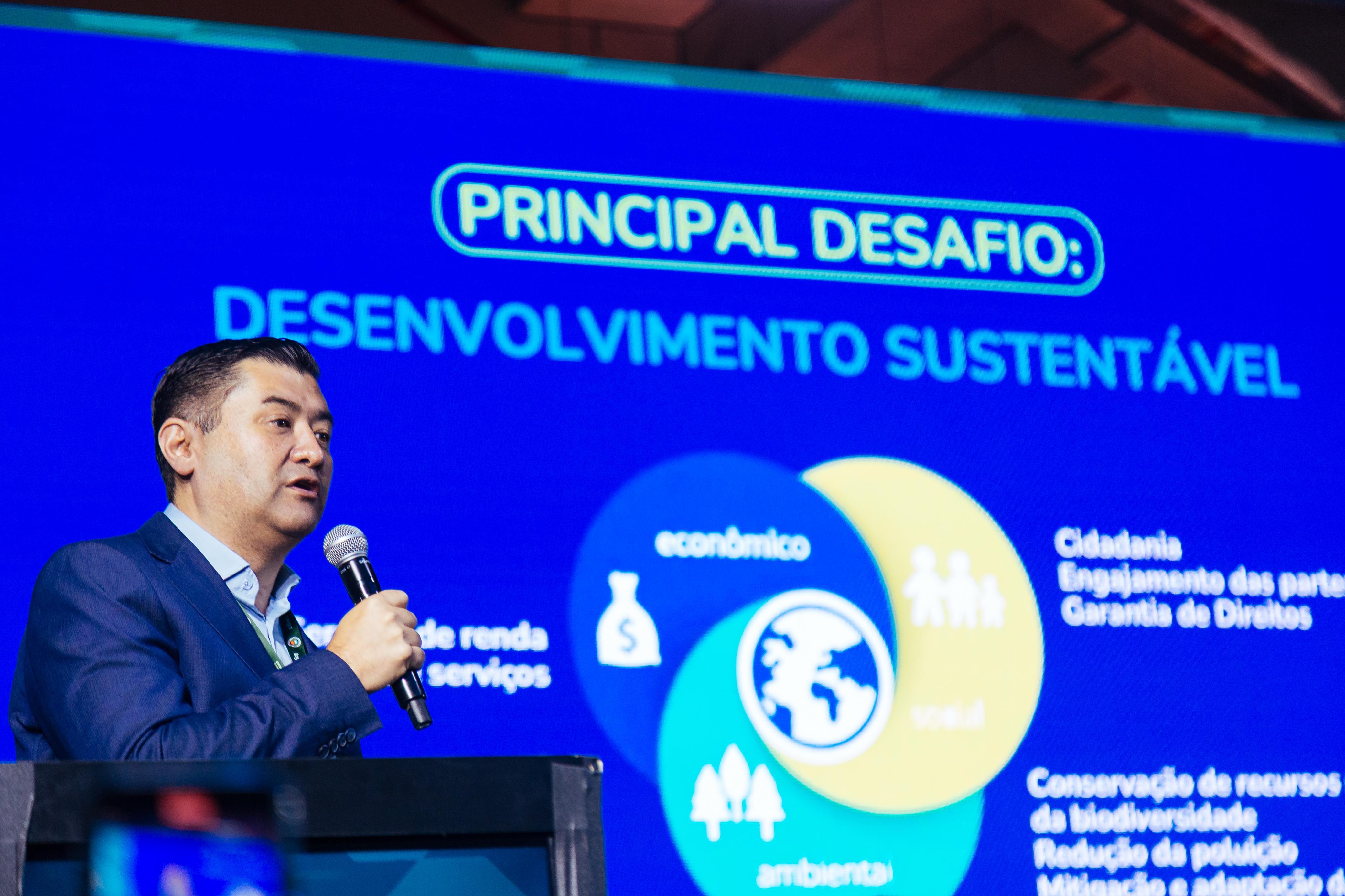 Homem de terno azul segura um microfone e fala em um palco durante apresentação sobre sustentabilidade. Ao fundo, telão azul exibe a frase “Principal desafio: Desenvolvimento sustentável” acompanhada de um diagrama com três áreas sobrepostas: econômico, social e ambiental.
