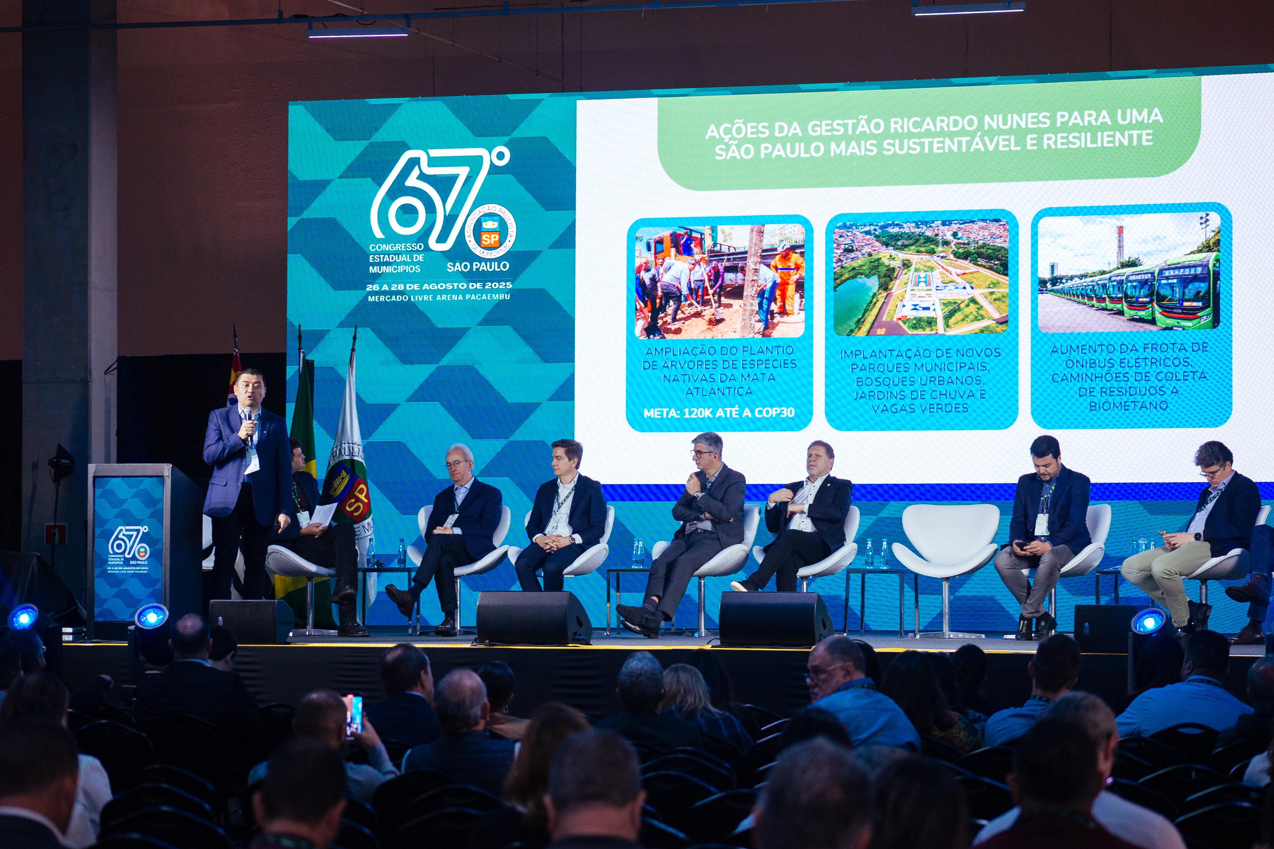 Auditório com plateia assistindo a uma mesa-redonda no 67º Congresso Estadual de Municípios em São Paulo. No palco, sete homens de terno estão sentados em cadeiras brancas, enquanto um deles fala ao microfone de pé. O telão exibe ações da gestão municipal relacionadas à sustentabilidade, como plantio de árvores nativas, implantação de novos parques e aumento da frota de ônibus elétricos.