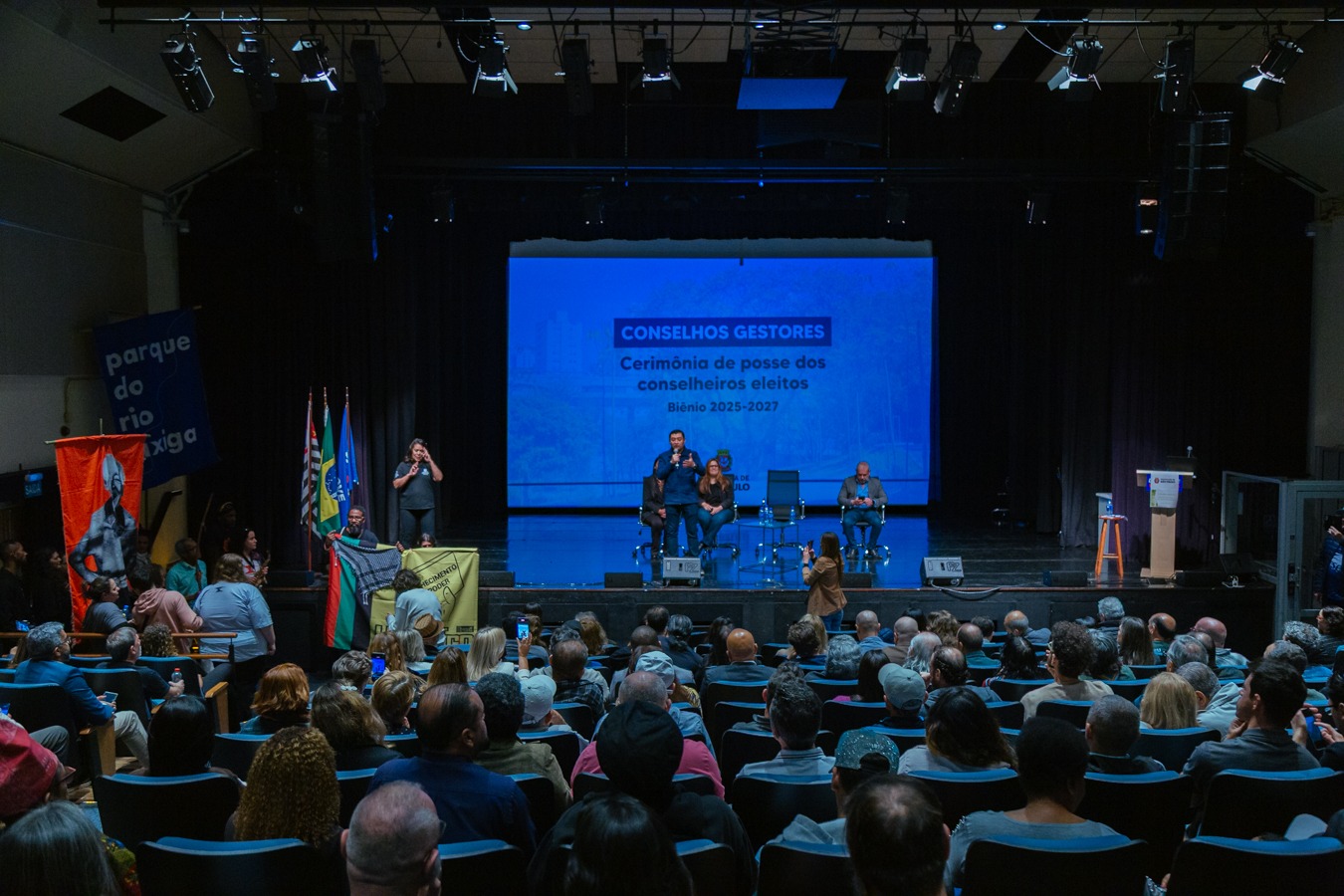 Cerimônia de posse dos conselheiros eleitos para o biênio 2025-2027, em um auditório cheio. No palco, há intérprete de Libras e representantes sentados. Bandeiras e cartazes de movimentos sociais compõem o cenário, reforçando a diversidade e a participação popular.