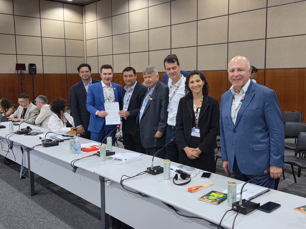 A imagem retrata uma cena de reunião ou conferência em uma sala moderna e bem iluminada. Em primeiro plano, há uma longa mesa de conferência escura, onde alguns participantes estão sentados, em sua maioria observando a frente da sala.  Em destaque, atrás da mesa, há um grupo de sete profissionais, predominantemente homens vestidos com ternos (cores escuras como preto, azul marinho e cinza). Eles estão em pé, posando para uma fotografia. O ambiente é profissional, com grandes janelas ou painéis de vidro ao fundo que refletem a luz, e a decoração da sala utiliza tons neutros como cinza, branco e madeira escura na mobília.
