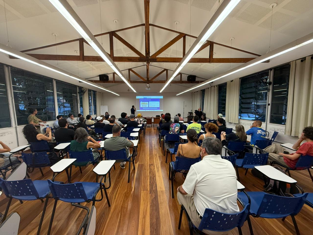 A imagem mostra uma sala ampla, com piso de madeira em tom marrom e brilho natural, e teto alto branco com vigas aparentes de madeira em tom castanho. O espaço é iluminado por longas luminárias lineares de luz branca, que acompanham o comprimento do teto. Diversas pessoas estão sentadas em cadeiras azuis com pranchetas acopladas, organizadas em fileiras voltadas para a frente da sala. O público veste roupas casuais em cores variadas, como preto, cinza, verde, azul e bege, com alguns detalhes em tons mais vivos. À frente, um telão branco exibe uma apresentação com fundo azul e elementos claros, posicionada acima de uma mesa de madeira escura. As paredes laterais são claras, em tons de branco e bege, com grandes janelas de vidro escuro e cortinas claras, criando contraste com o piso e o mobiliário.