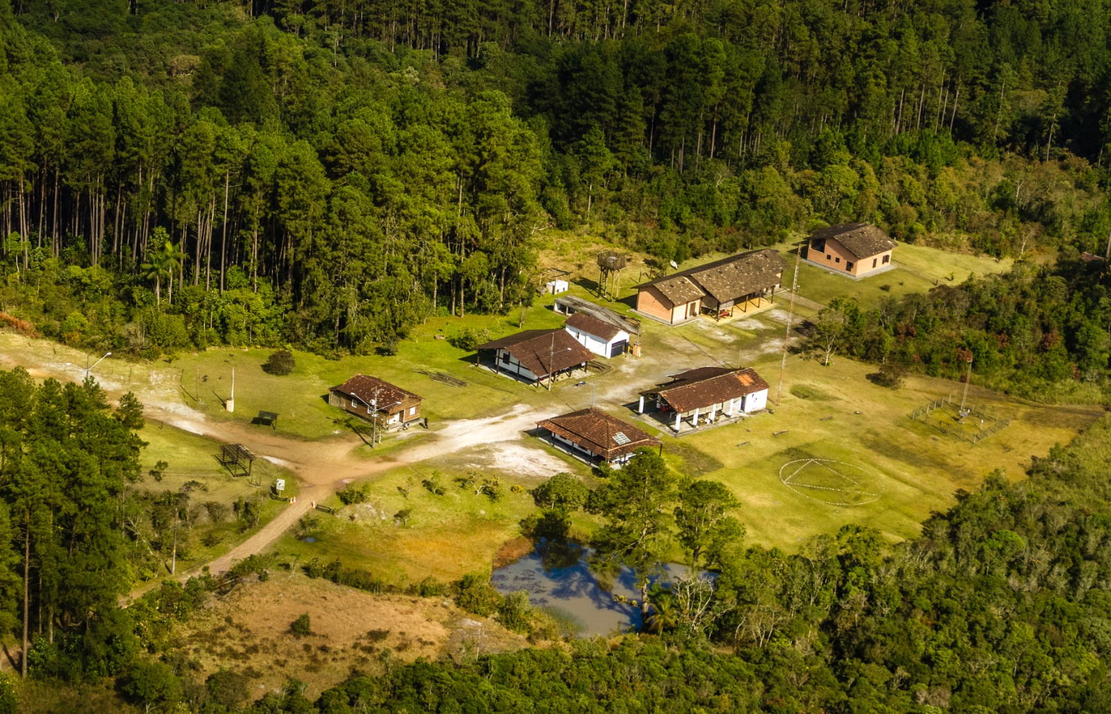 Imagem aérea em plano aberto mostrando um pequeno conjunto de construções em meio a uma área extensa de natureza. A maior parte da imagem é ocupada por vegetação densa, com muitas árvores altas de copas cheias em tons variados de verde, do verde-escuro ao verde mais claro e amarelado.  No centro da cena, há uma clareira ampla com gramado em tons de verde claro e áreas de terra batida em bege e marrom claro, formando caminhos que ligam os prédios. Espalhadas por essa clareira, aparecem várias construções térreas de tamanho médio e pequeno, com telhados de cerâmica em tom marrom-avermelhado e paredes em cores claras, como branco e bege, além de algumas em tom mais terroso. As edificações estão organizadas de forma espaçada, criando a impressão de um pequeno núcleo ou vila rural.  À direita da área central, vê-se um espaço aberto gramado com marcações claras no chão, formando um desenho circular ou oval, em tom branco ou muito claro, sobre o verde da grama. Na parte inferior da imagem, há um pequeno lago ou açude, com água escura refletindo o verde das árvores ao redor, criando manchas de azul-esverdeado e marrom.  O contraste entre o verde intenso da floresta ao redor, os tons claros dos caminhos de terra e o marrom dos telhados destaca o conjunto de construções no meio da paisagem natural, transmitindo uma sensação de isolamento, tranquilidade e integração com o ambiente.
