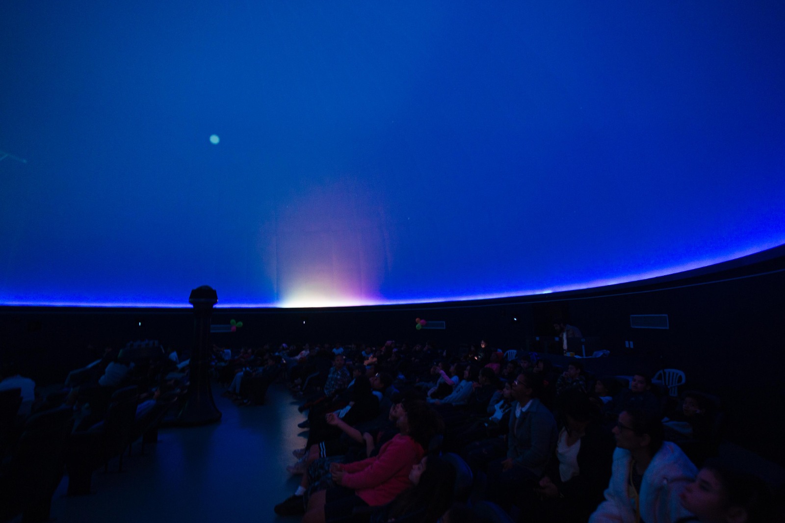 Público sentado em um planetário, observando projeção azul no teto em formato de cúpula.