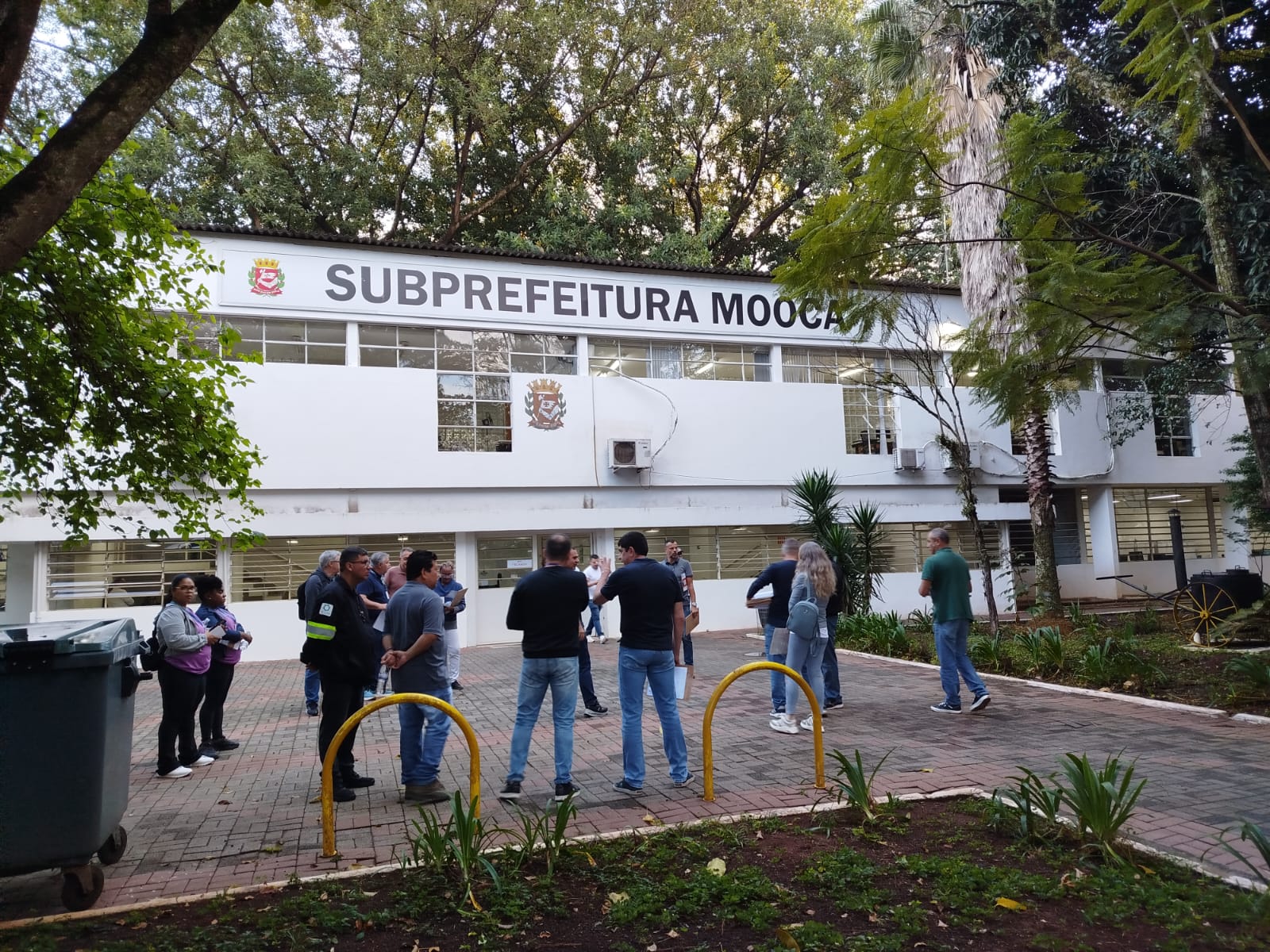 Fachada da Subprefeitura Mooca, com um grupo de pessoas reunido na área externa do prédio, cercada por árvores e jardins.