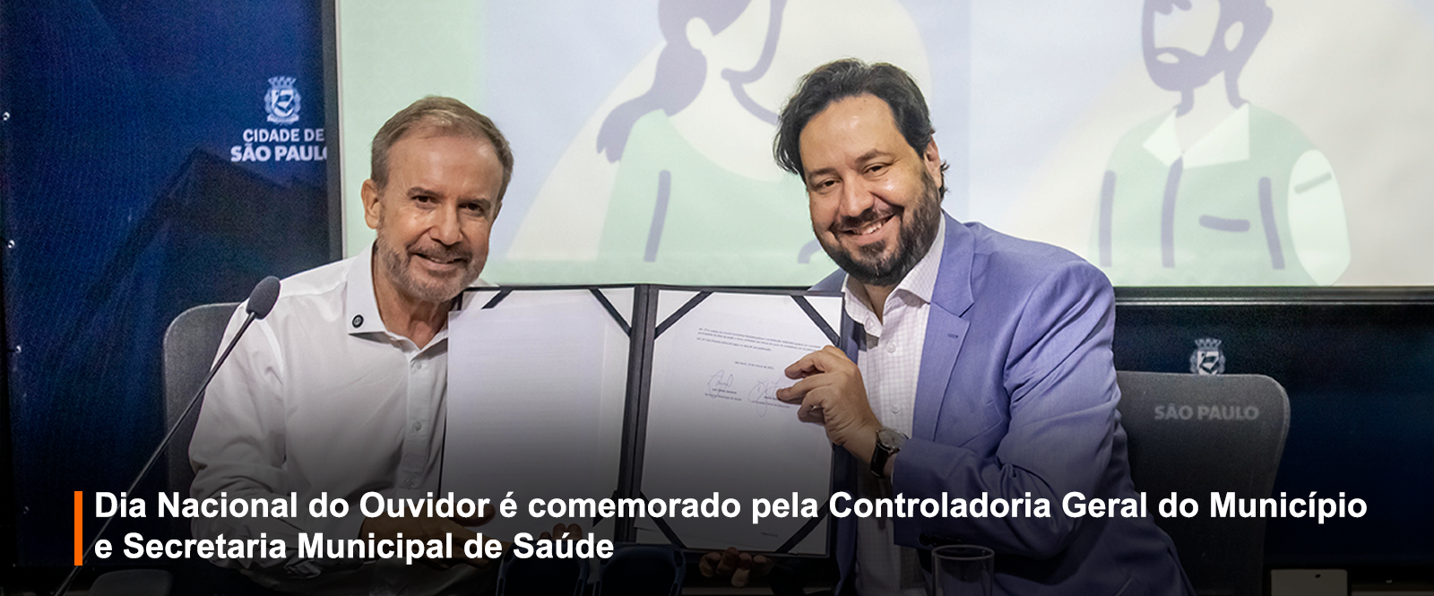 Fotografia tirada durante um evento institucional. Dois homens estão sentados lado a lado ao centro da imagem, sorrindo e segurando um documento aberto com duas páginas visíveis. À esquerda, o homem veste camisa branca. À direita, o homem usa blazer azul claro e camisa social branca. Ambos estão com semblantes felizes e voltados para a câmera. Ao fundo, uma tela exibe ilustrações de personagens estilizados, e à esquerda está o brasão da Cidade de São Paulo com a inscrição 