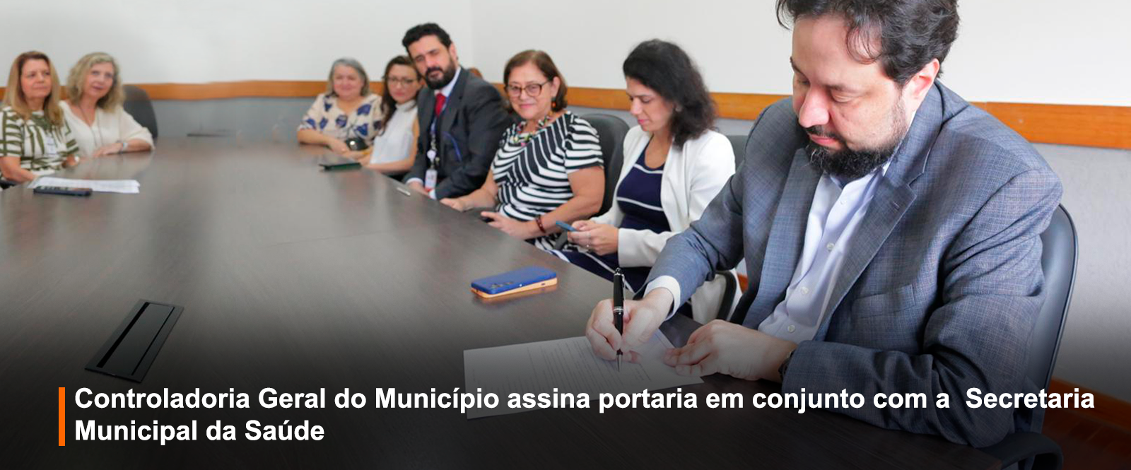 #TextoDescritivo #ParaTodosVerem  Fotografia colorida tirada em uma sala de reuniões. À direita da imagem, um homem de terno cinza assina um documento com uma caneta preta sobre a mesa. Ele está concentrado, com o corpo inclinado para frente. No lado esquerdo, vemos uma fileira de pessoas sentadas ao redor da mesma mesa, todas voltadas para a câmera. São nove pessoas ao todo, entre mulheres e homens, com expressões neutras ou levemente sorridentes. Algumas seguram celulares ou papéis. A mesa é grande e de madeira escura, e o ambiente possui paredes claras com faixa de madeira ao fundo.  Na parte inferior da imagem, sobre uma faixa preta translúcida, está o texto em letras brancas e negrito: “Controladoria Geral do Município assina portaria em conjunto com a Secretaria Municipal da Saúde”. Uma barra vertical laranja aparece ao lado esquerdo da palavra “Controladoria”, funcionando como elemento gráfico de destaque.