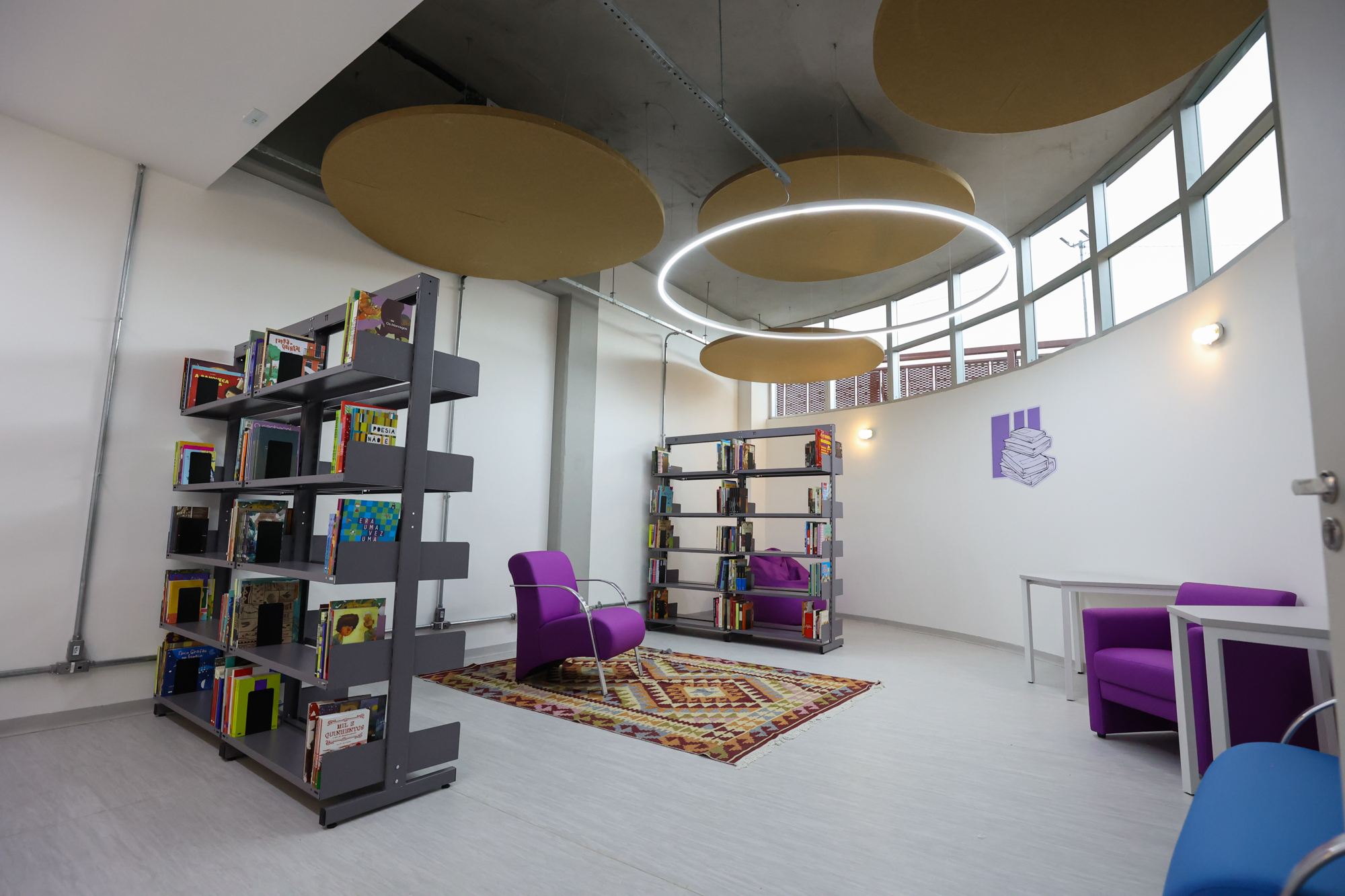 Imagem da biblioteca do Centro TEA. Nela há mesas, cadeiras, armários, estante com livros e tapete.