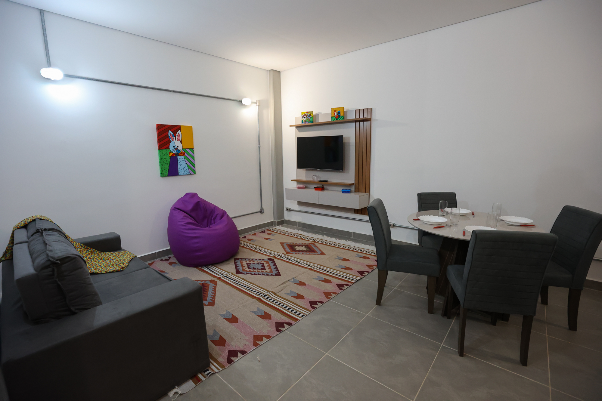 Imagem da sala do Vida Diária do Centro TEA. Na sala há um sofá, tapete, puff, um quadro de coelho na parede, televisão, cadeiras e uma mesa com pratos, copos e talheres.
