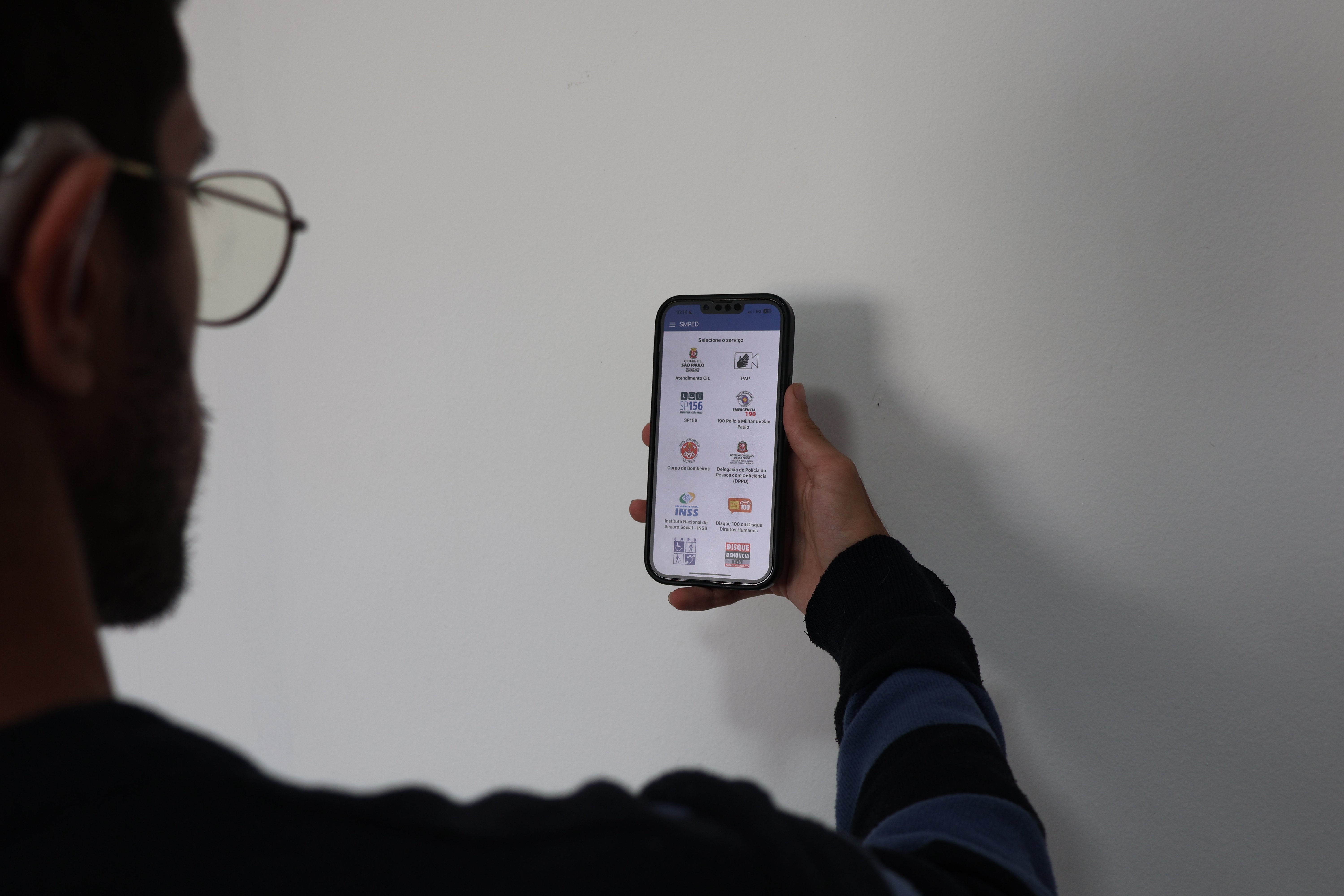 Um homem que usa aparelho auditivo está de costas olhando para o celular que está em sua mão. Na tela está a página principal do aplicativo CIL-MPED com diversos ícones de serviços.