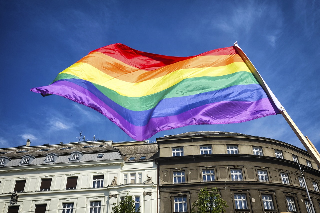 A imagem exibe uma bandeira do orgulho LGBTQIA+ ondulando sob um céu azul claro. A bandeira possui listras horizontais nas cores vermelho, laranja, amarelo, verde, azul e roxo, simbolizando diversidade e inclusão. Ao fundo, há uma construção arquitetônica com varandas e janelas, em cores neutras, que destaca a bandeira em primeiro plano. A luz do sol ilumina a cena, criando uma sensação de celebração e visibilidade. A imagem transmite um forte sentimento de orgulho e apoio à comunidade LGBTQIA+.