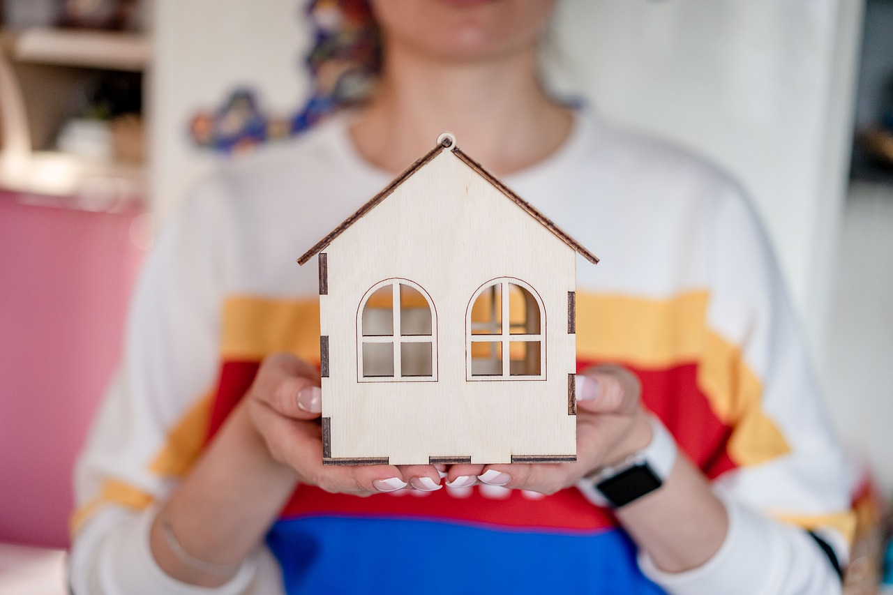 A imagem mostra uma pessoa segurando uma pequena maquete de uma casa de madeira. A casa tem um design simples, com um telhado acentuado e janelas em formato arredondado. A pessoa está em primeiro plano, com as mãos na frente, destacando a maquete. Ela usa uma blusa de cores vivas, com listras horizontais em vermelho, azul e amarelo. O fundo é desfocado, mas parece ser um ambiente bem iluminado, com uma parede de cor clara e alguns objetos não identificáveis. A imagem transmite um sentimento de criatividade e aconchego.