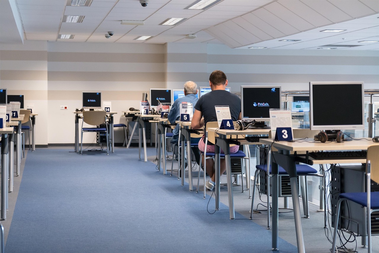 A imagem mostra um ambiente moderno com várias estações de computador. Existem mesas dispostas em linha, cada uma com um computador de tela plana e fones de ouvido, identificadas por números. Três pessoas estão sentadas, de costas, em diferentes posições. O piso é azul e as paredes têm listras claras, criando um espaço organizado e iluminado, sugerindo um local de aprendizado ou trabalho.