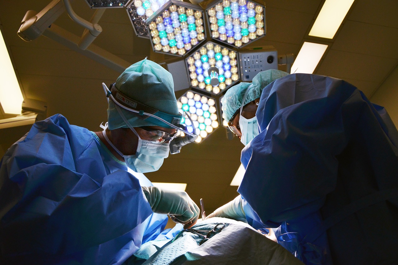 A imagem mostra um ambiente cirúrgico com dois dentistas em primeiro plano. Eles estão vestidos com roupas cirúrgicas azuis, incluindo toucas, máscaras e óculos de proteção. Os cirurgiões estão concentrados em um procedimento, com um dos profissionais utilizando um instrumento cirúrgico. O fundo é iluminado por um complexo sistema de luzes cirúrgicas em formato hexagonal, que emitem uma luz clara e brilhante. 