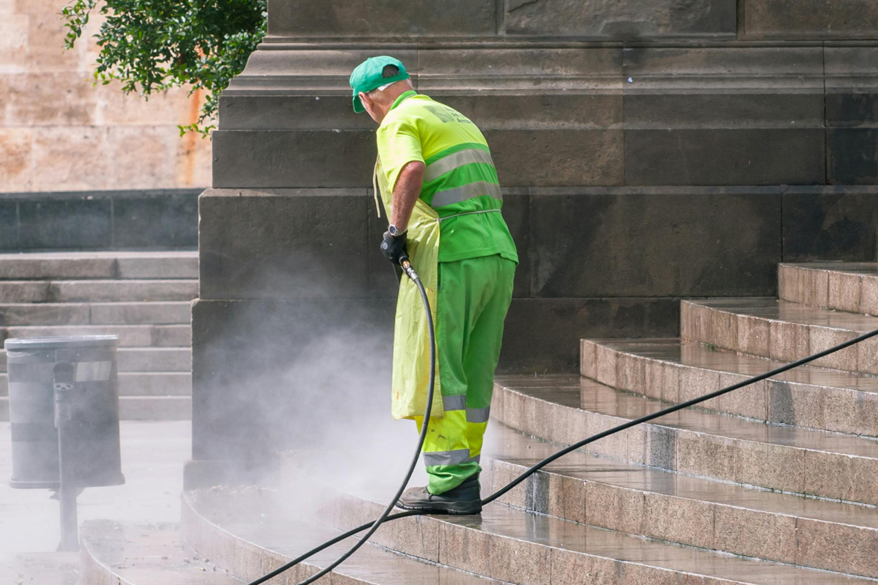 A imagem mostra um trabalhador de limpeza usando um uniforme amarelo fluorescente com faixas reflexivas, realizando a lavagem de escadas de pedra. Ele está de costas, segurando uma mangueira que emite água sob pressão, criando um jato de spray e uma leve névoa. Ao lado, há uma lixeira cinza. O ambiente está ao ar livre, com a luz do sol iluminando a cena e algumas árvores ao fundo. As etapas da escada estão molhadas, refletindo a ação de limpeza em curso. A imagem transmite um senso de trabalho e cuidado com o espaço público.
