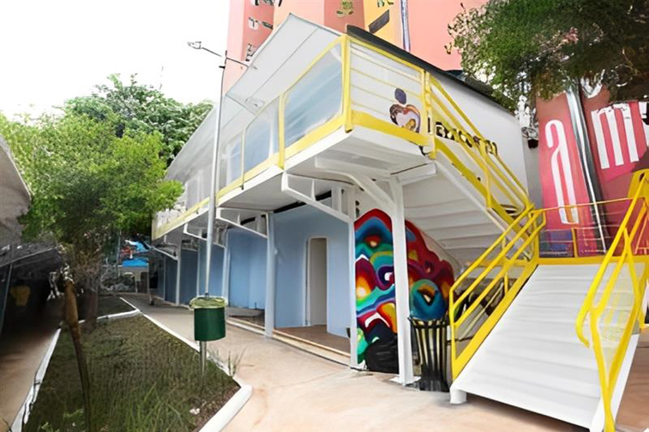 A imagem apresenta um edifício de dois andares com uma estrutura moderna. O primeiro andar é acessível por uma rampa com corrimões amarelos. O edifício tem paredes de cor azul com decorações artísticas coloridas em uma das laterais. Há uma área externa com gramados e árvores, proporcionando um ambiente natural. O céu é claro, e a iluminação parece ser boa. O espaço parece ser bem projetado para a acessibilidade e está situado em uma área urbana.