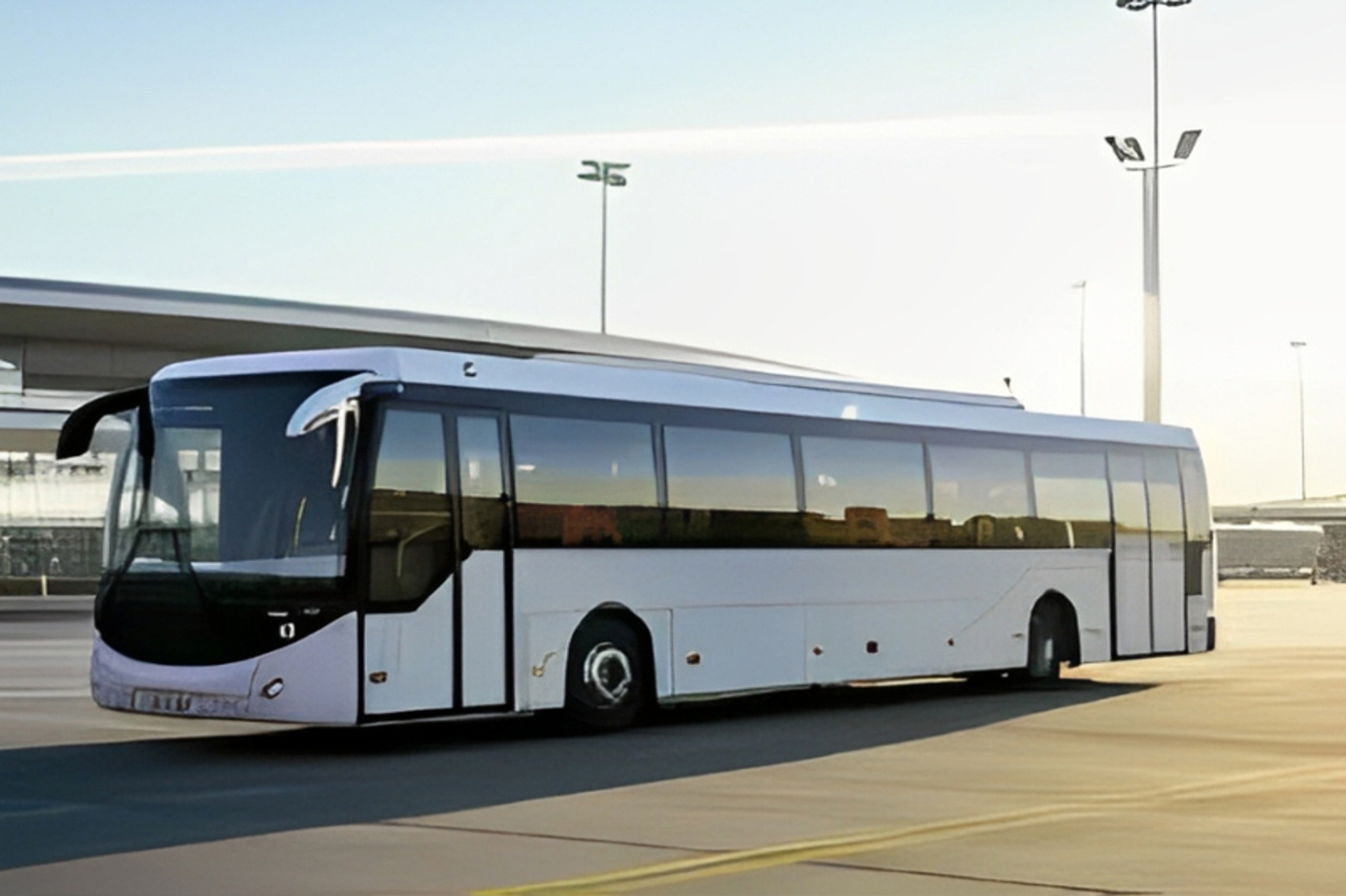 A imagem mostra um ônibus moderno estacionado em um terminal, rodeado por um ambiente amplo e ensolarado. O ônibus é predominantemente branco, com janelas grandes e faixas pretas ao redor. Ele possui uma estrutura limpa e aerodinâmica, com uma parte frontal arredondada e uma porta de entrada na lateral. Ao fundo, pode-se ver postes de iluminação e uma pista ampla, sugerindo um espaço de transporte, como um aeroporto ou terminal rodoviário. A iluminação natural cria uma atmosfera clara e aberta, destacando o veículo em um dia ensolarado.