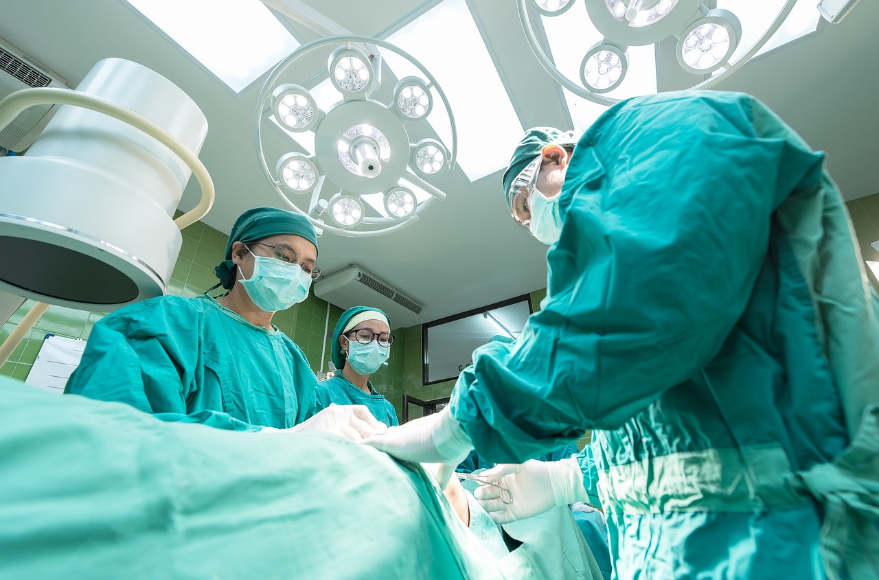 A imagem retrata uma cena de cirurgia em um ambiente hospitalar. Três profissionais de saúde estão vestidos com roupas cirúrgicas verdes, incluindo toucas e máscaras. Dois deles estão próximos, concentrados em um procedimento, enquanto um terceiro observa. No centro da imagem, pode-se ver a parte superior de um paciente coberta por um lençol cirúrgico. Acima, há um sistema de luz cirúrgica com várias fontes de luz brilhante, proporcionando iluminação ao campo cirúrgico. O fundo é composto por paredes azuis e verdes, criando um ambiente clínico e limpo. A cena transmite um ambiente de cuidado e profissionalismo na área de saúde.