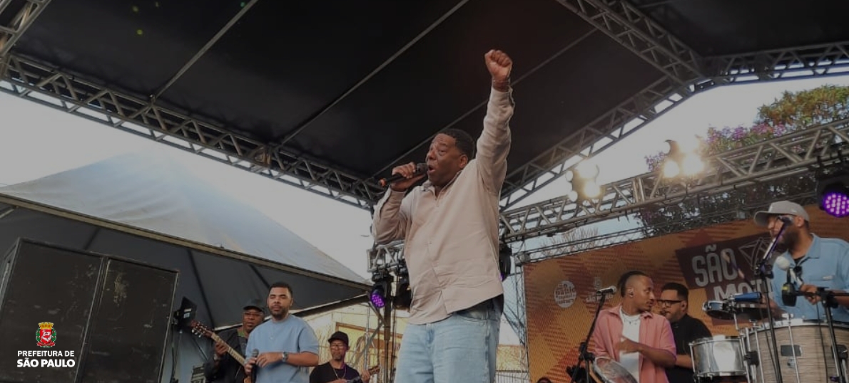 O cantor Netinho de Paula no centro do palco, vestindo camisa bege e calça jeans clara, segura o microfone e levanta o braço direito em gesto de celebração