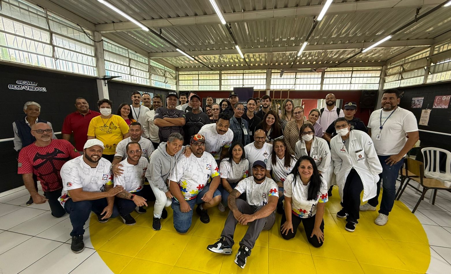 #PraTodosVerem: Espaço iluminado, paredes pretas com janelas, o chão branco com um grande círculo amarelo pintado. Cerca de 40 pessoas reunidas, a maioria utilizando camisetas na cor branca ou preta, e sorrindo para a foto. No canto esquerdo, duas pessoas vestindo camiseta vermelha e uma pessoa de amarelo.